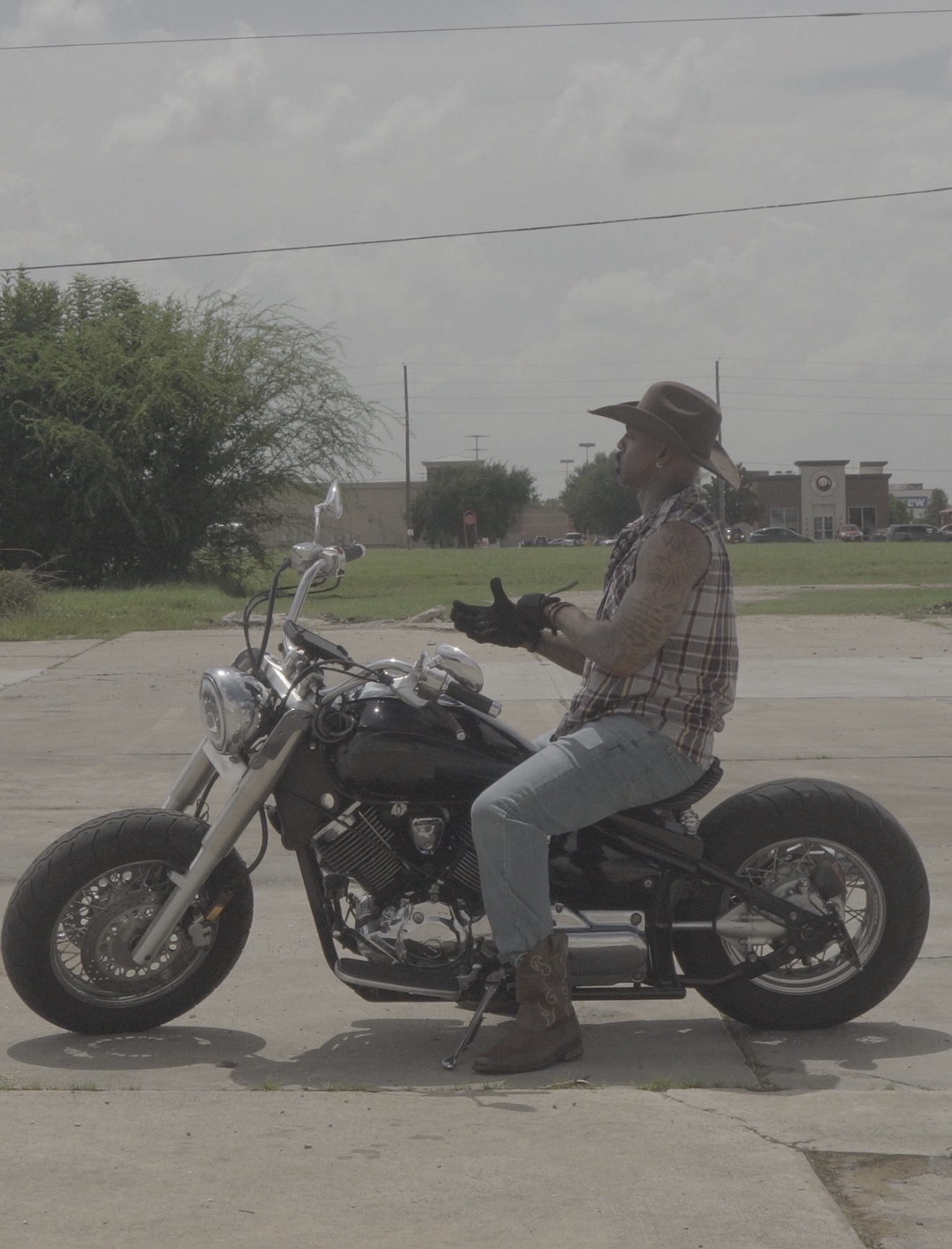 Person sitting on a motorcycle in an outdoor parking lot, wearing a hat, plaid shirt, and jeans, holding a phone.