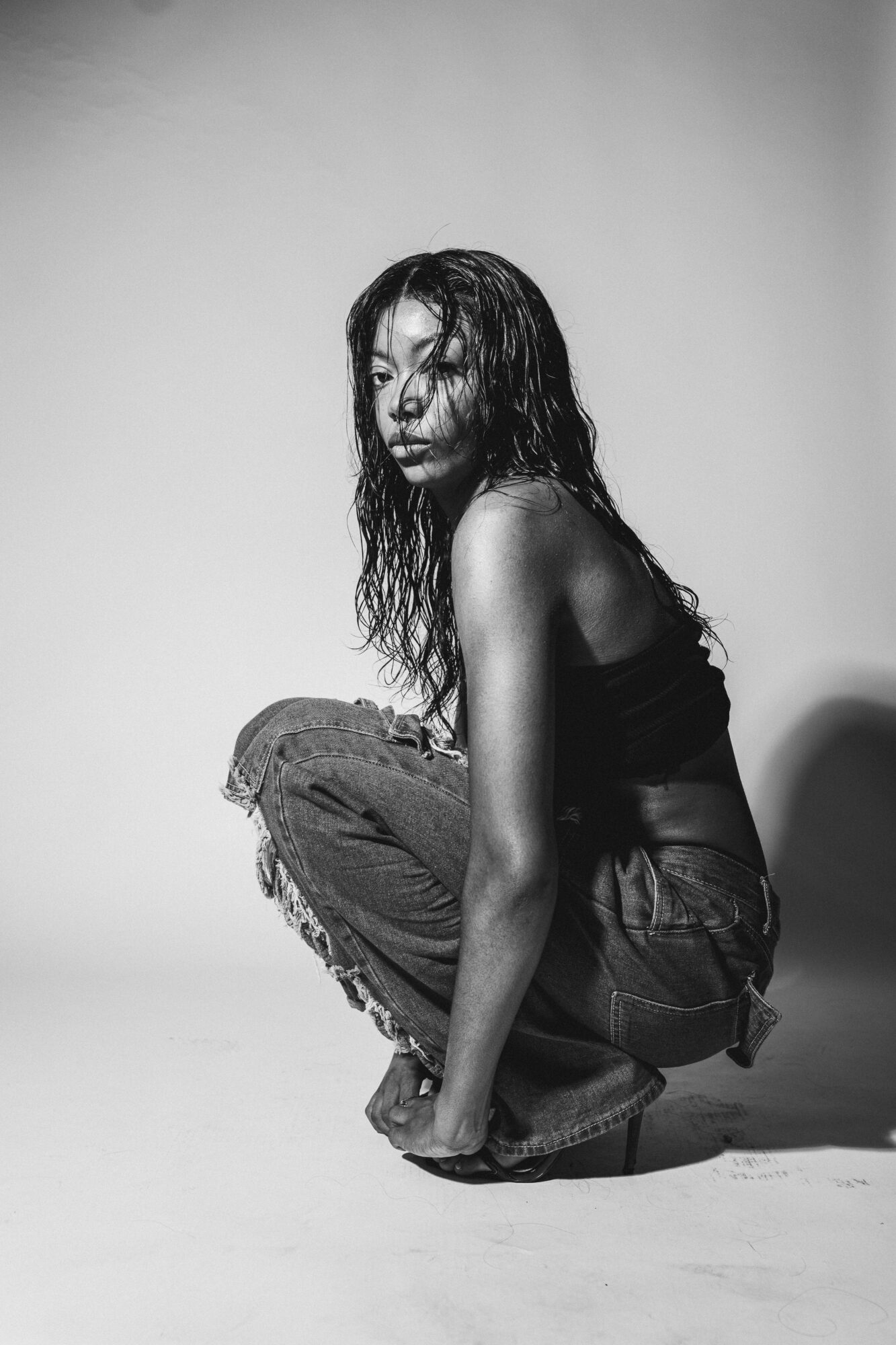 Young woman with curly hair crouching, wearing a tank top and jeans, in a studio setting.