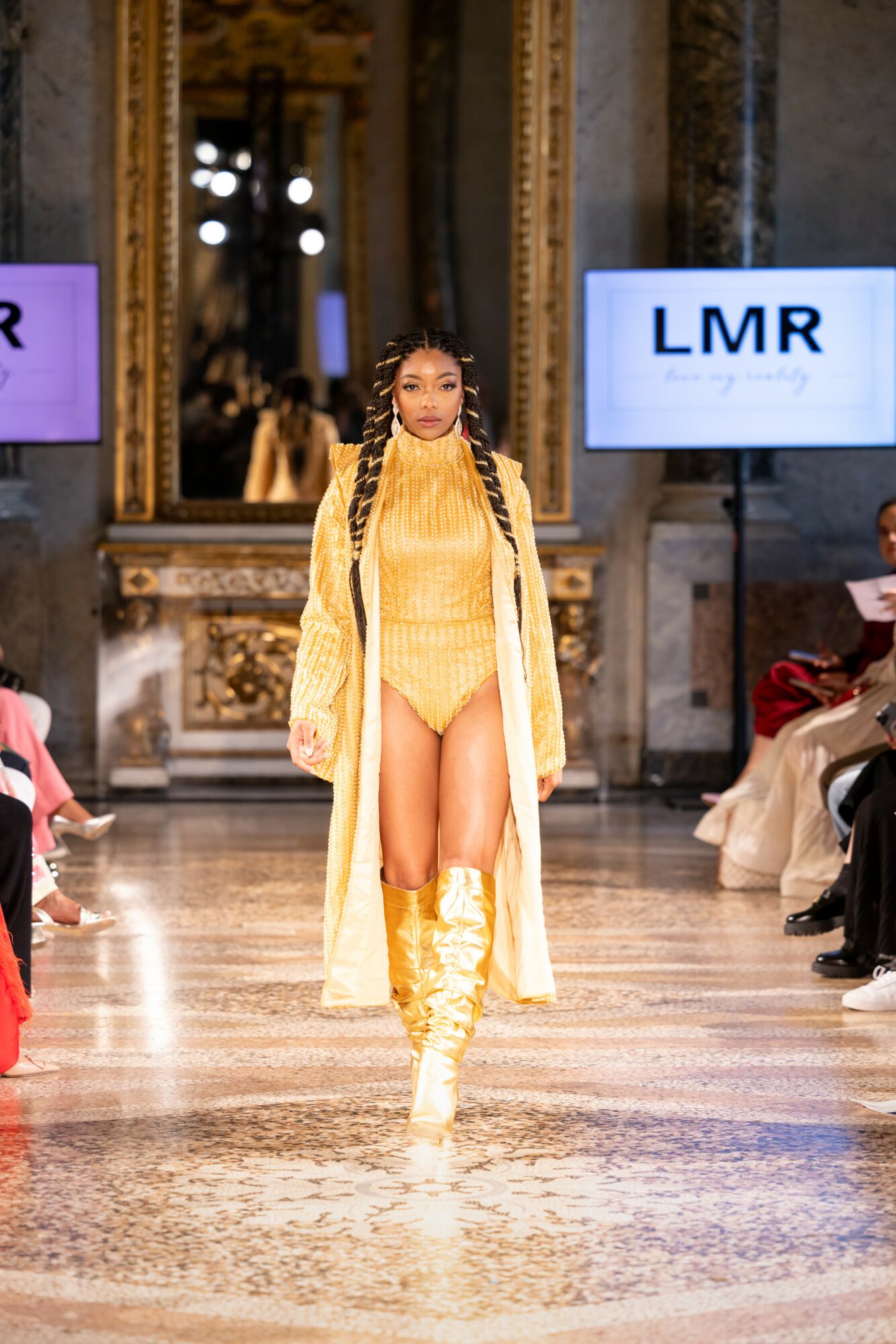 Model walking on runway in yellow outfit with long coat and thigh-high boots, in ornate room with mirrors and screens