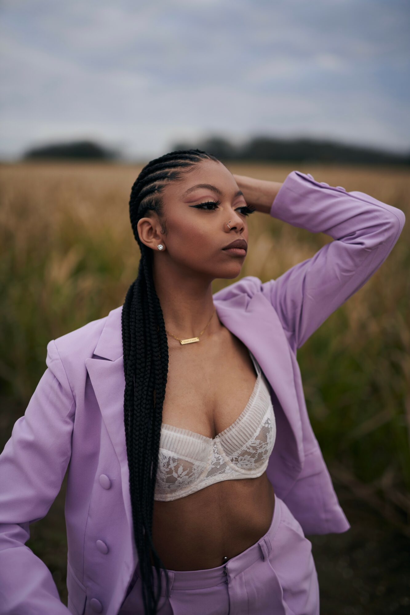 Woman with braided hair in a field, wearing a lavender blazer and white lace top, looking to the side outdoors.