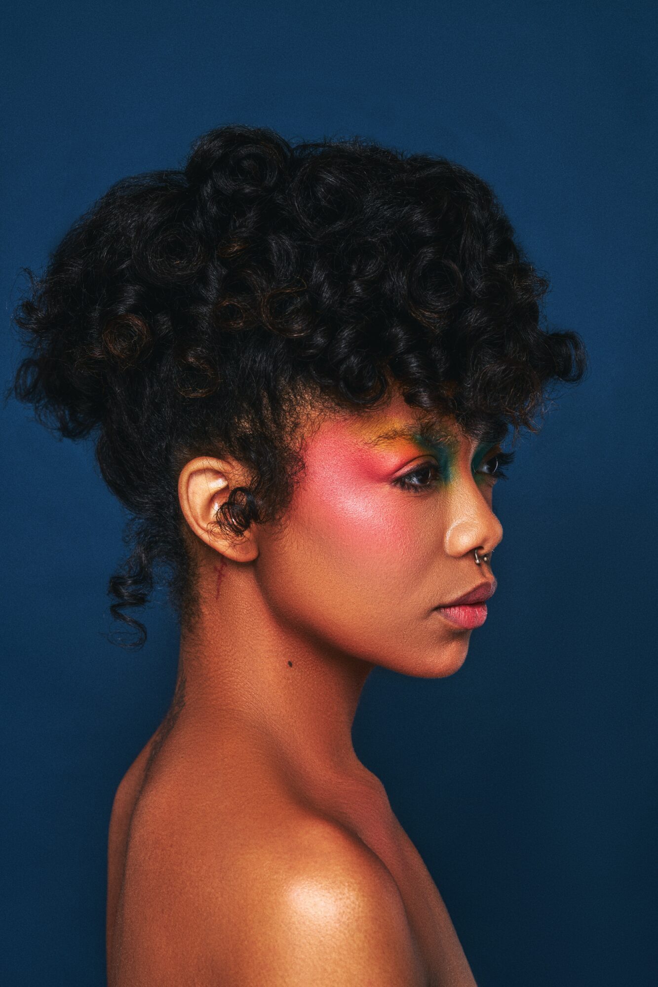 Profile of a woman with curly hair and colorful makeup against a dark blue background.