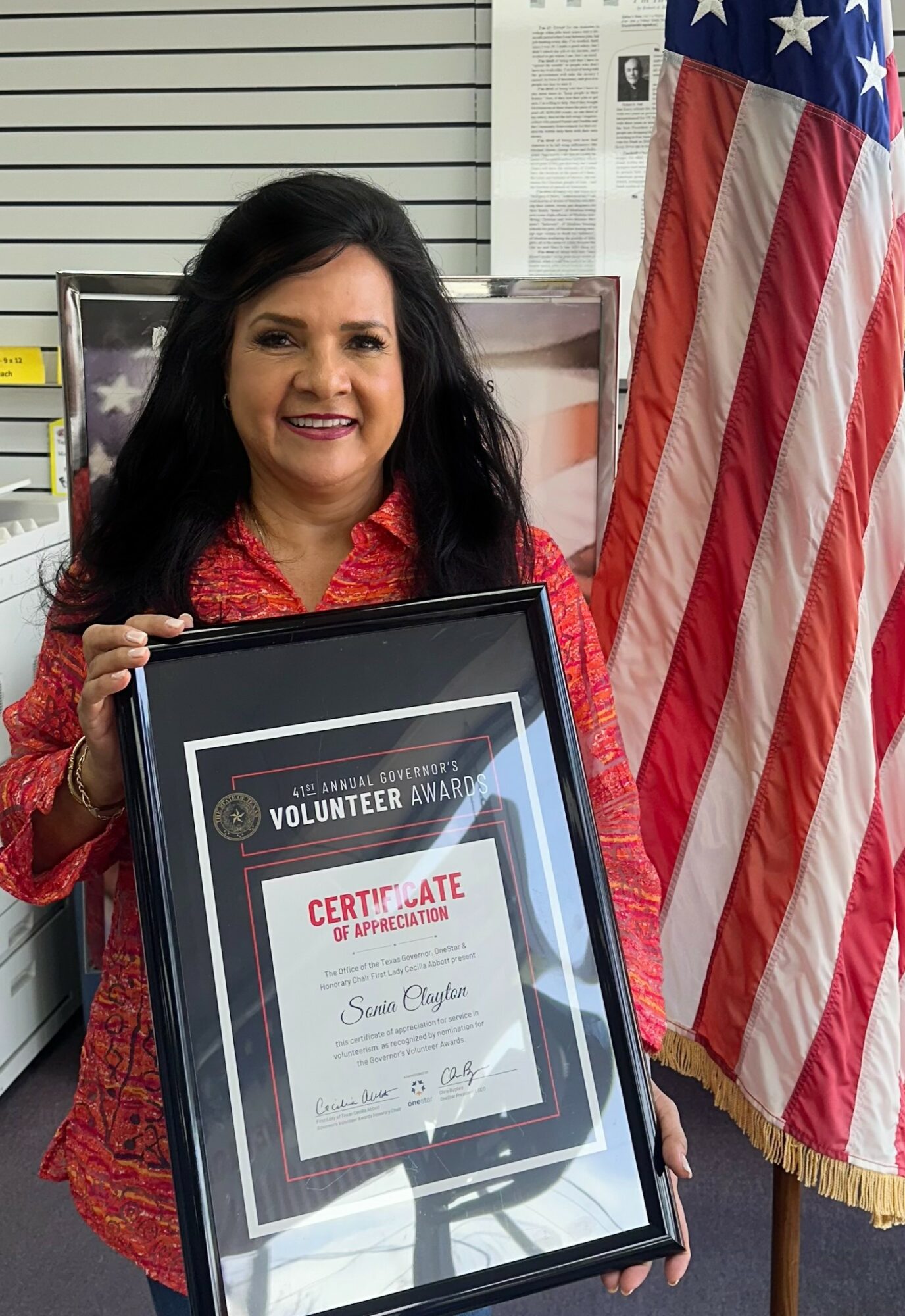 Woman holding a framed certificate, standing next to an American flag, smiling, in an indoor setting.