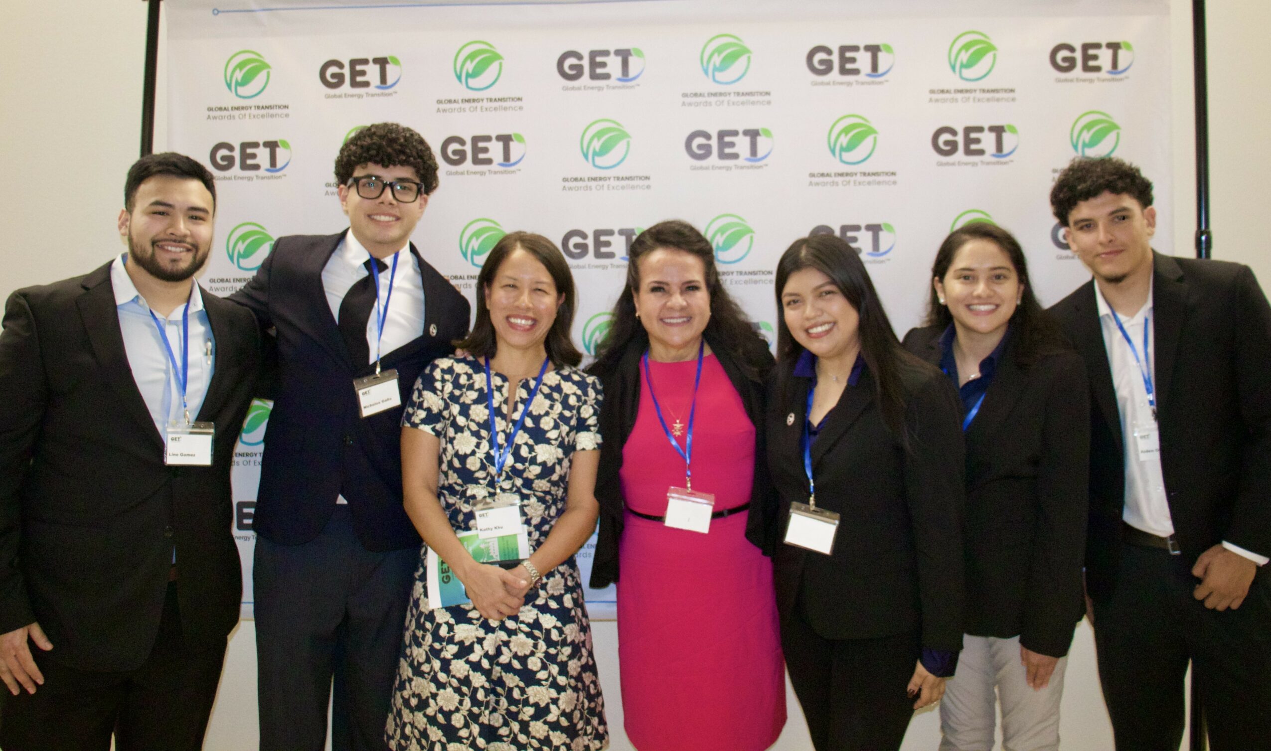 Group of seven people standing in front of a GET banner, smiling, wearing conference badges, and formal attire.