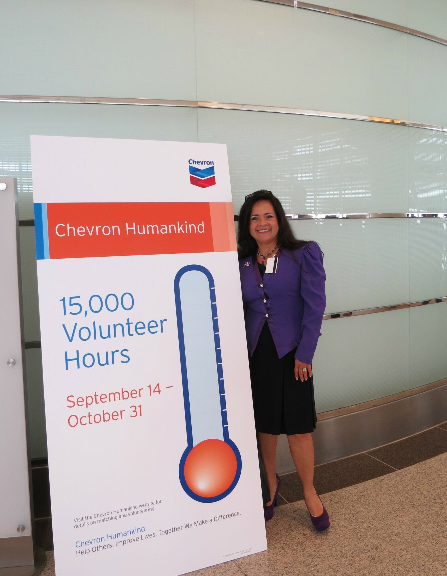 Woman standing next to a Chevron Humankind sign with volunteer hours and dates, smiling, in an indoor setting.