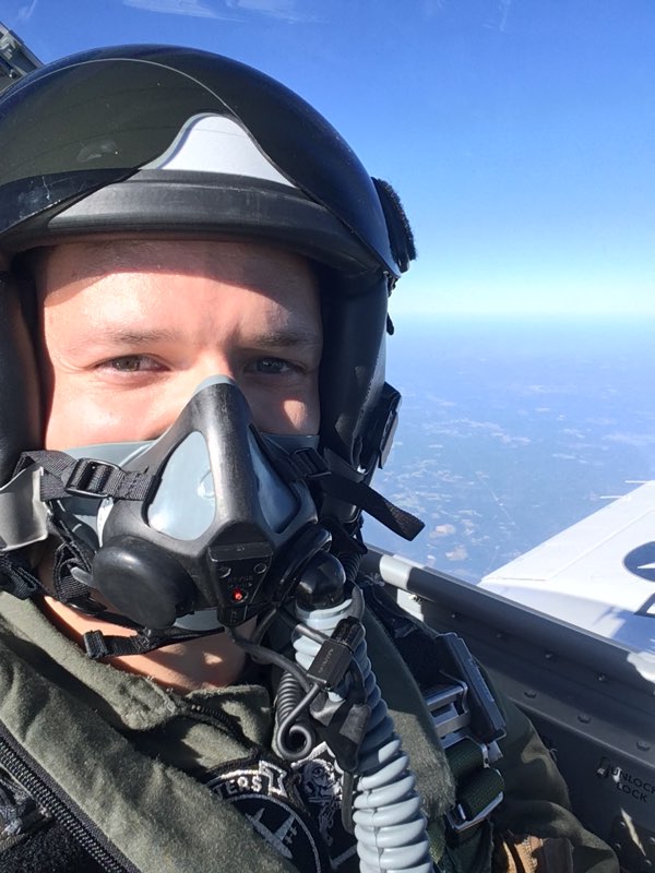 Person wearing a helmet and oxygen mask inside an aircraft cockpit, with sky and clouds outside.