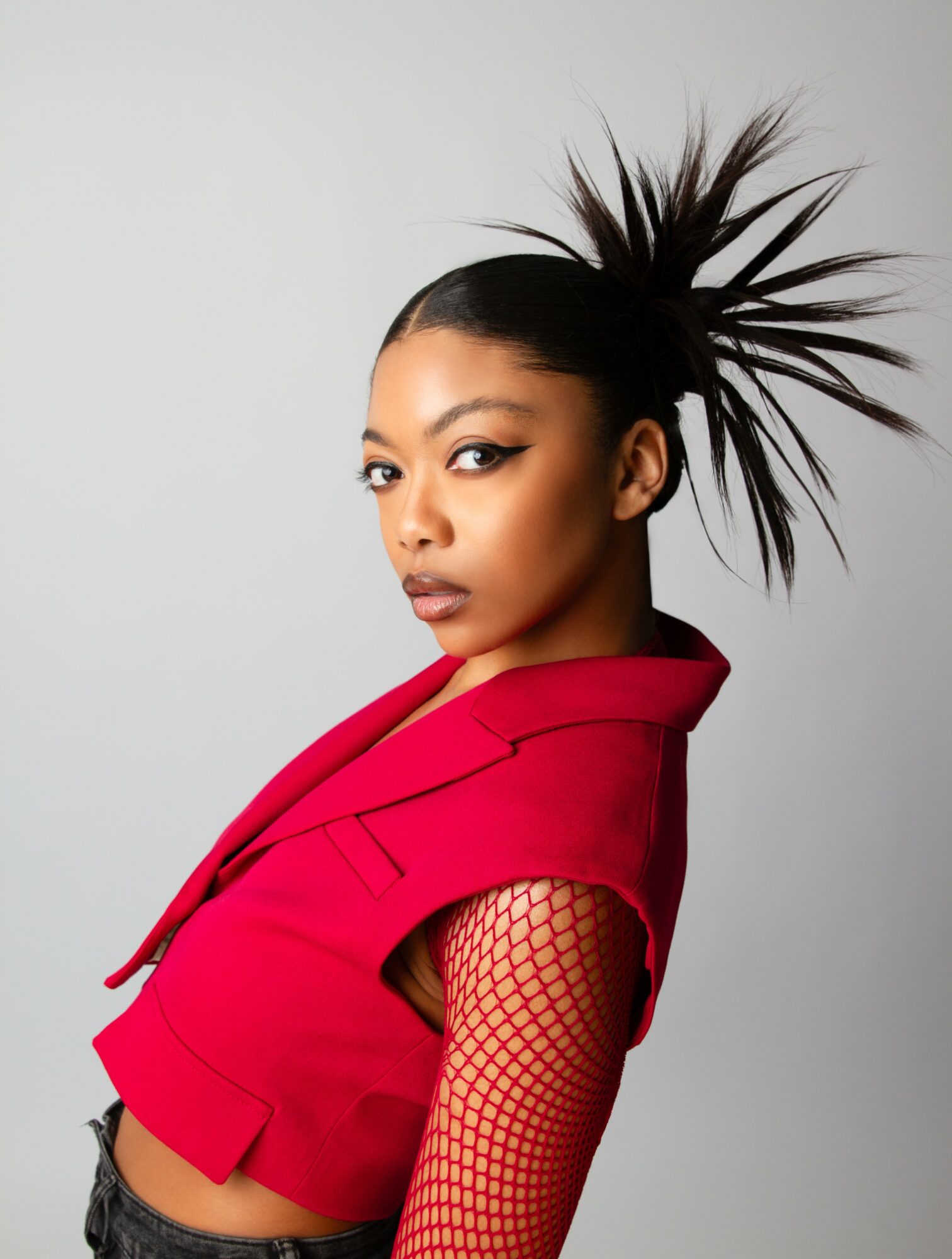 Young woman with spiked hairstyle wearing a red jacket and fishnet sleeve, looking at the camera against a plain background.