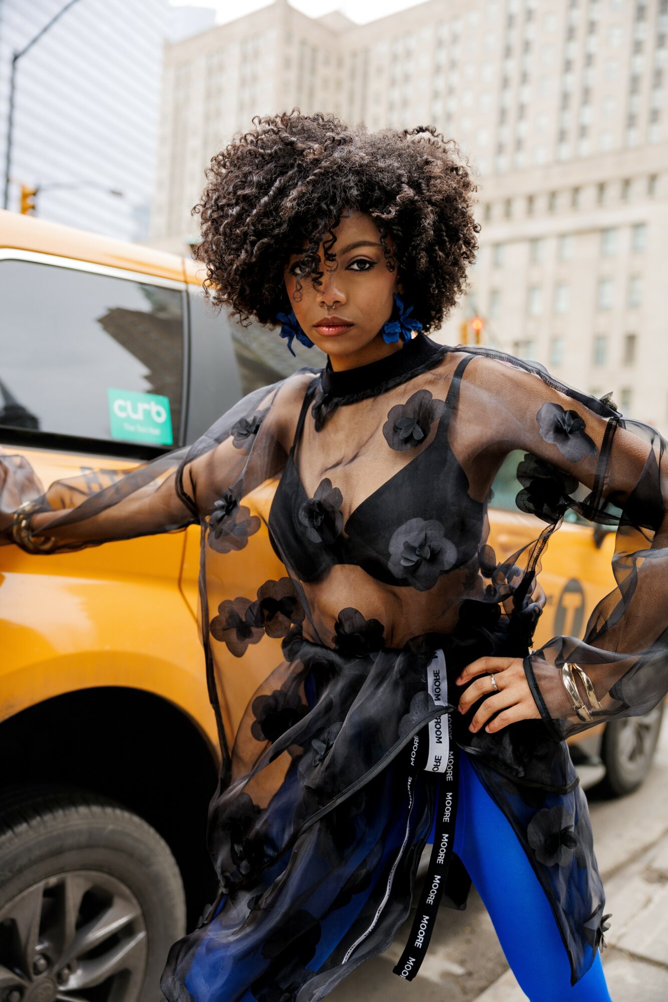 Woman with curly hair in a sheer black top with floral patterns, standing in front of a yellow taxi in an urban setting.