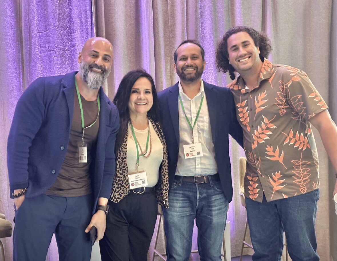 Group of four people smiling, standing in front of purple and beige curtains, wearing conference badges and casual clothing.
