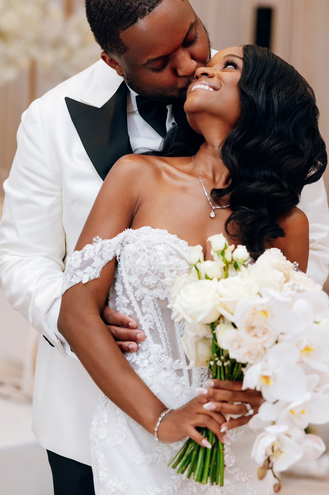 Couple in wedding attire, the man kissing the woman on her forehead, she holds a bouquet of white flowers, smiling.