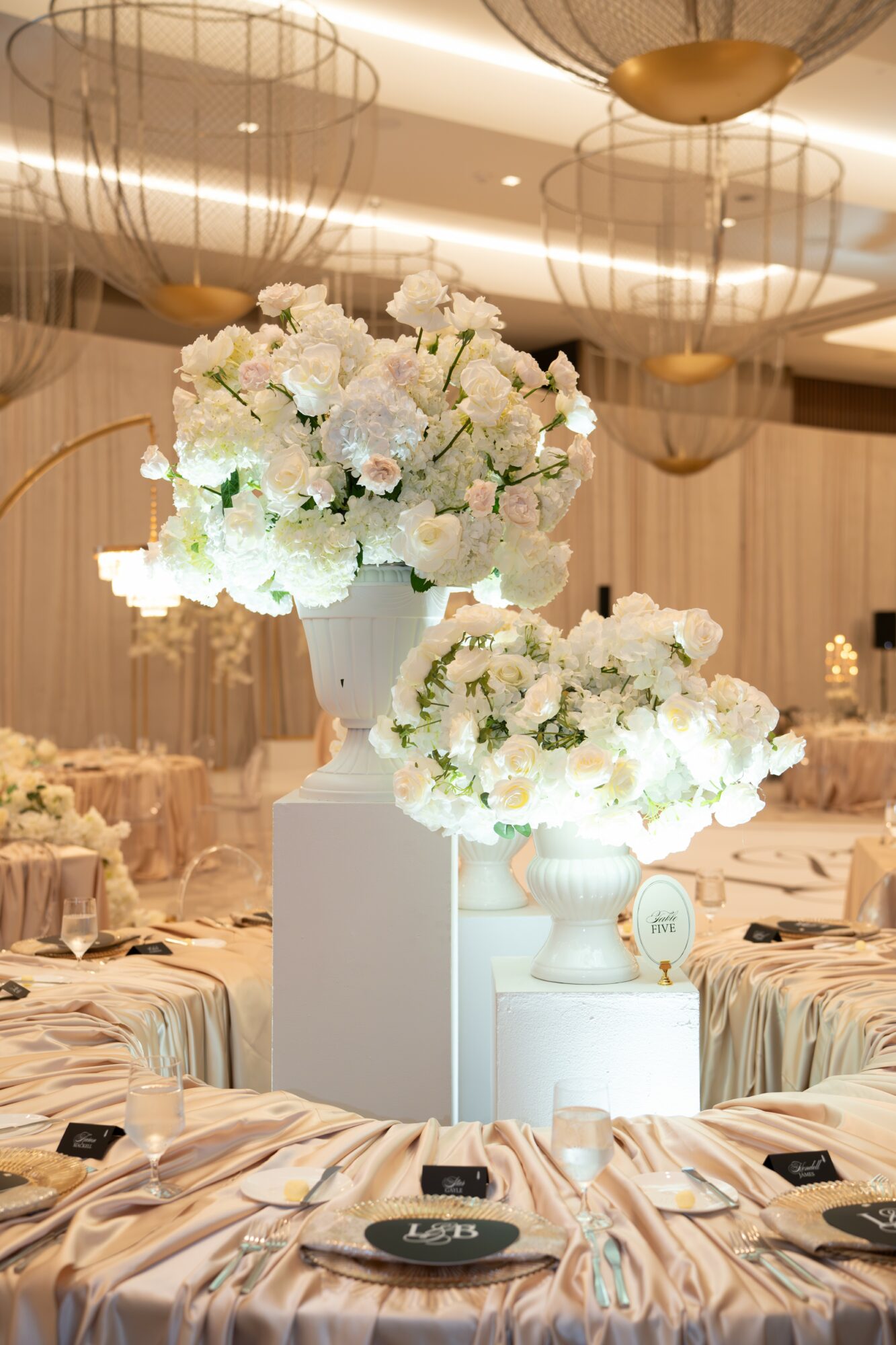 Elegant event space with large white floral arrangements on white pedestals, round tables with beige tablecloths, and warm lighting.