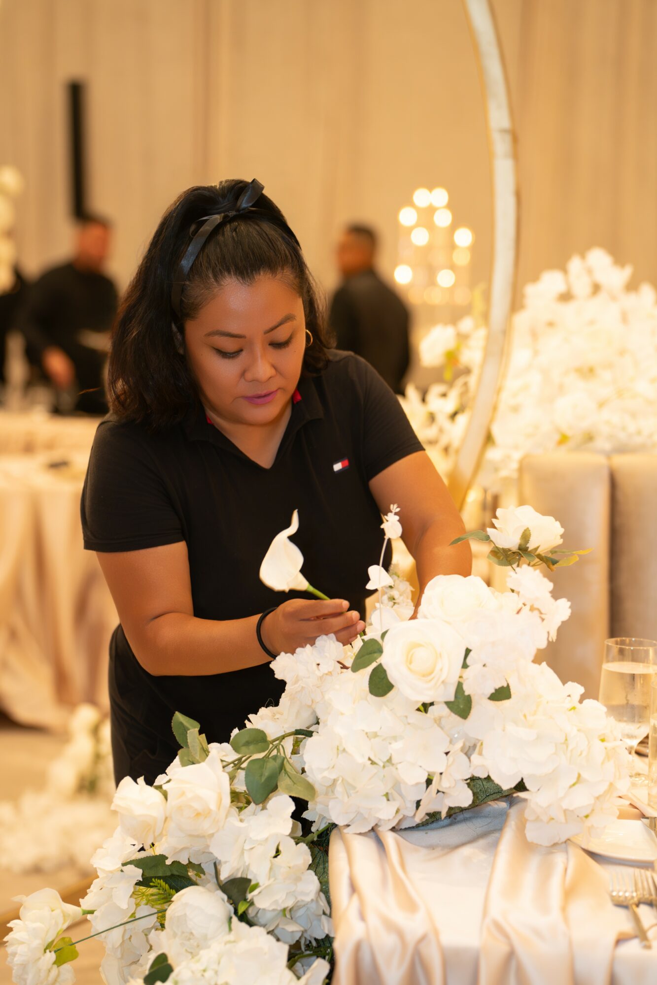 Woman arranging white flowers on a table at an event, with blurred background and warm lighting.