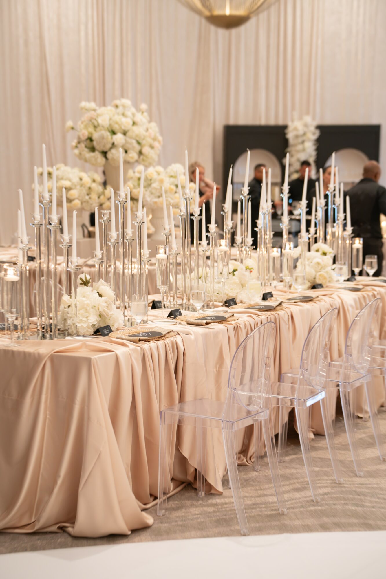 Elegant table with floral centerpieces, candles, and draped tablecloths at a formal event, with chairs and a backdrop in the background.