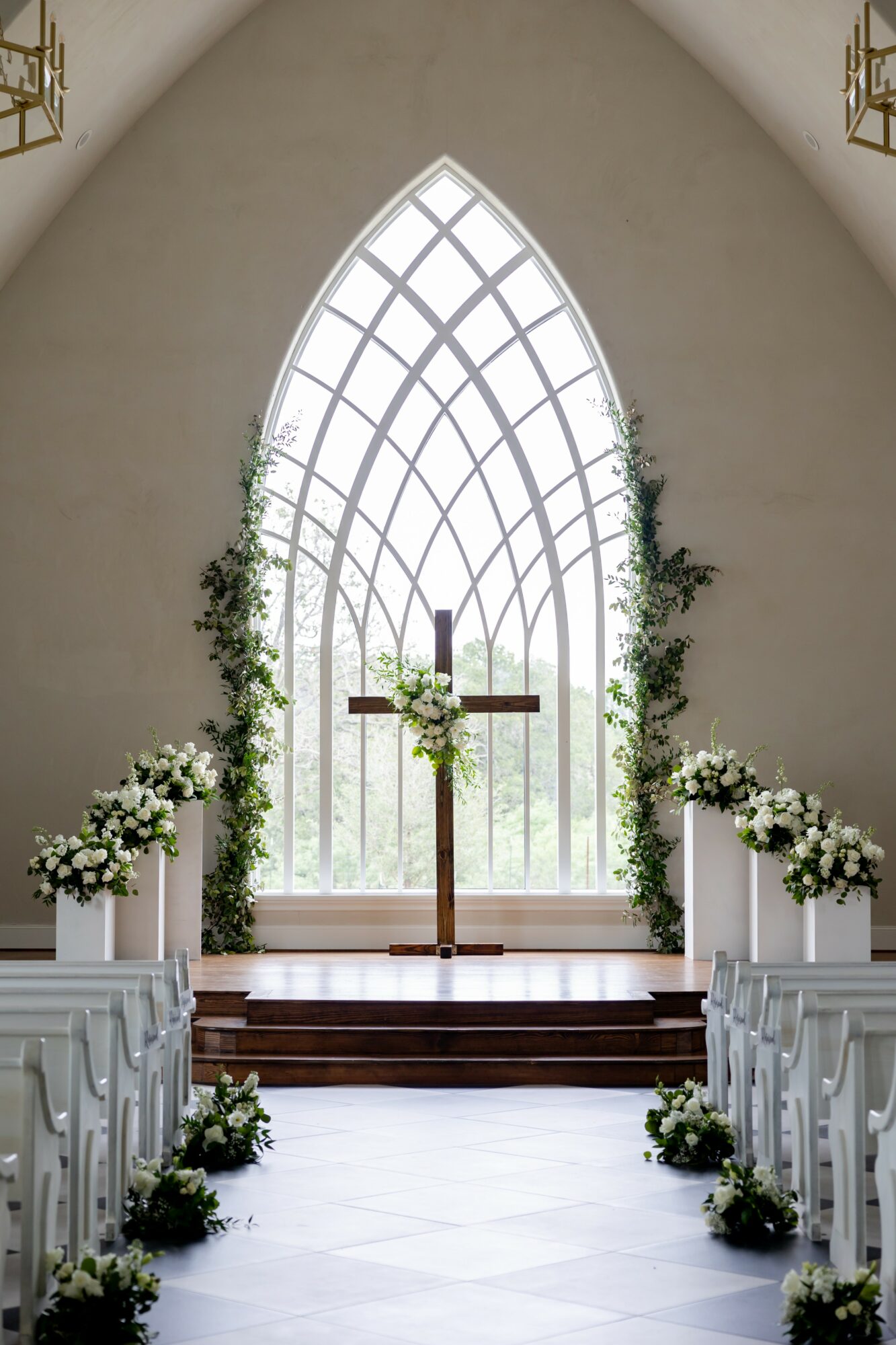Interior of a church altar with a cross, floral arrangements, and a large arched window behind.