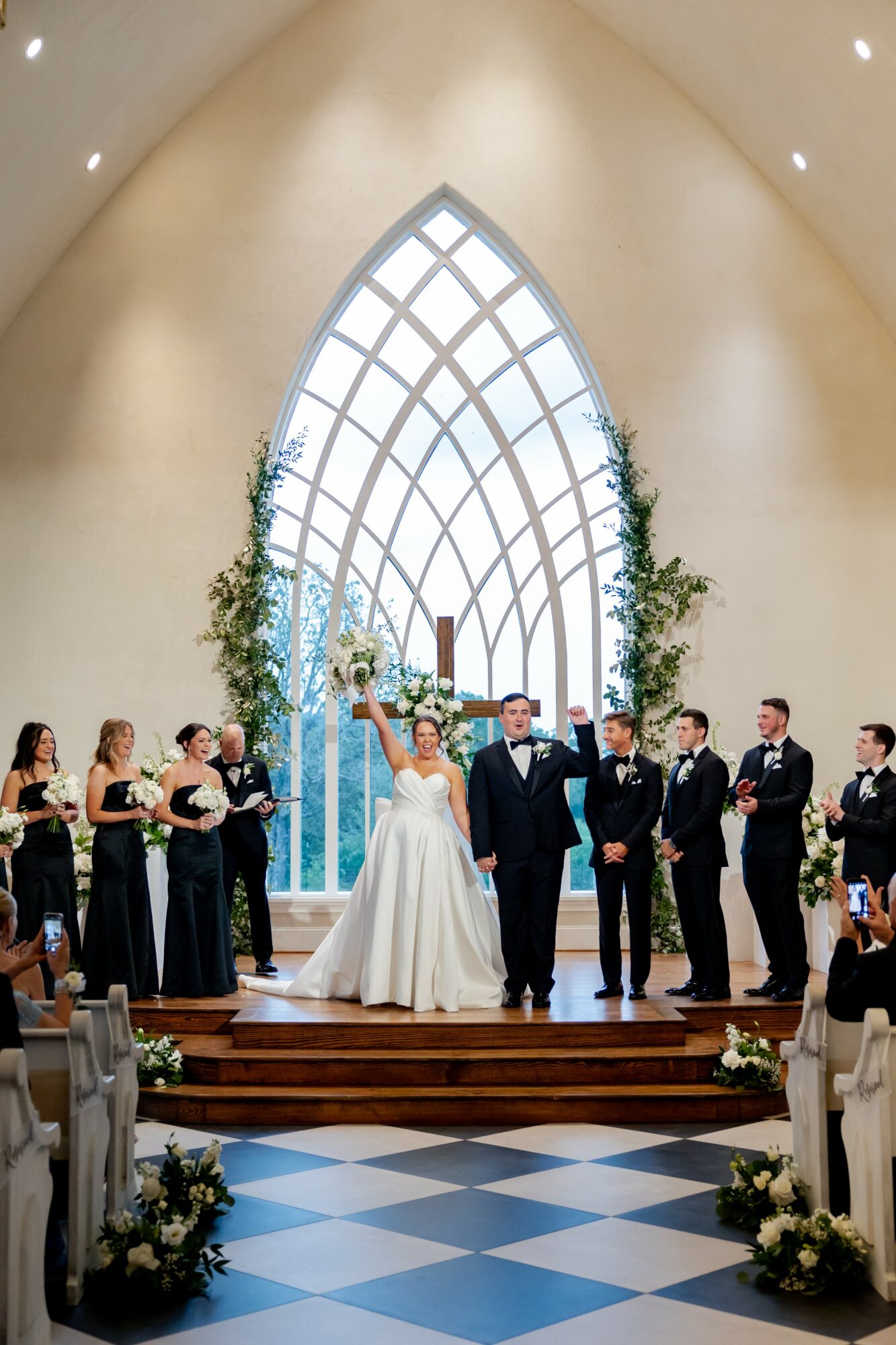 Wedding ceremony with bride and groom standing in front of a large arched window, surrounded by bridesmaids and groomsmen, inside a church.