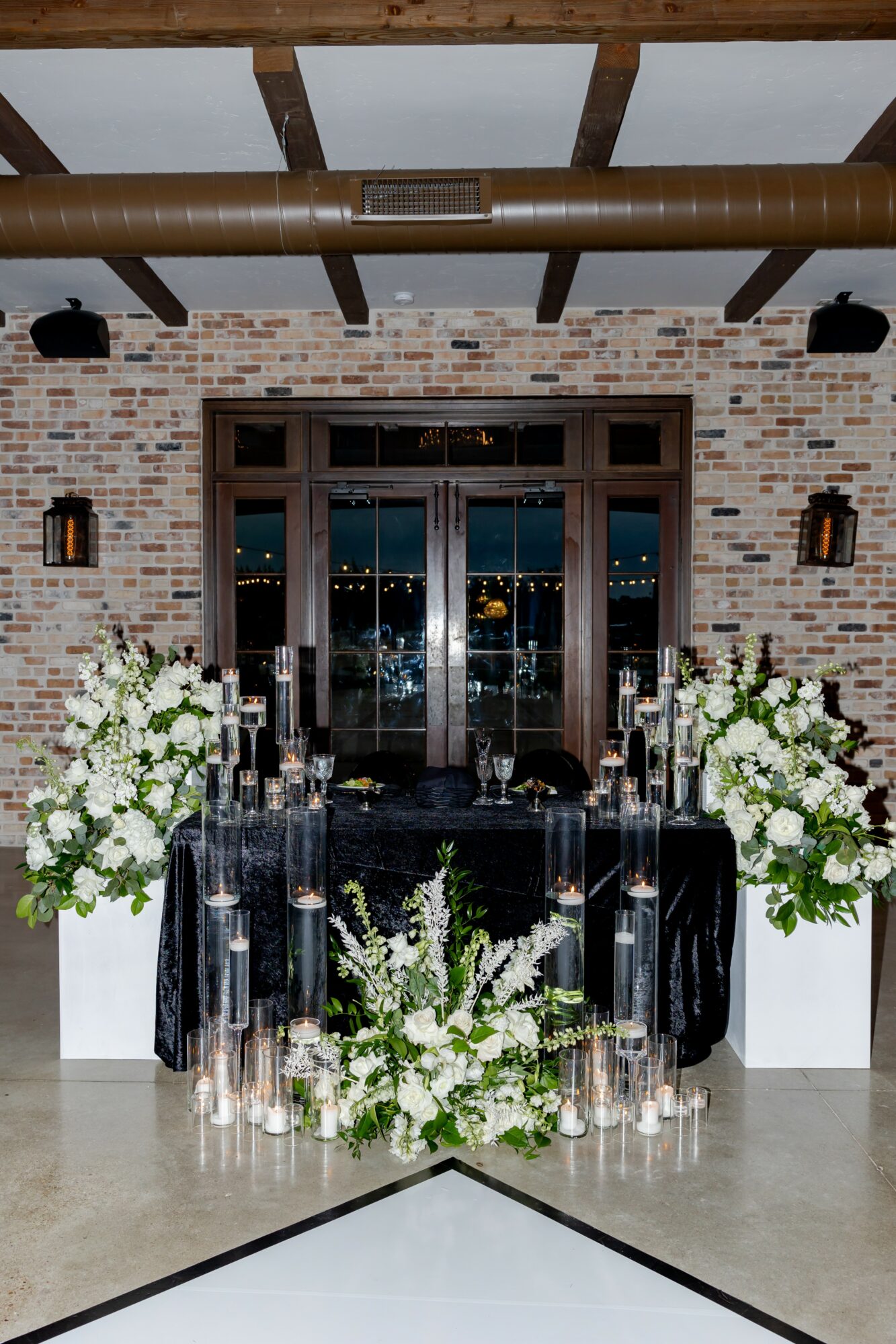 Decorated table with floral arrangements and candles in front of large glass doors, brick wall background, ceiling beams, and wall sconces.