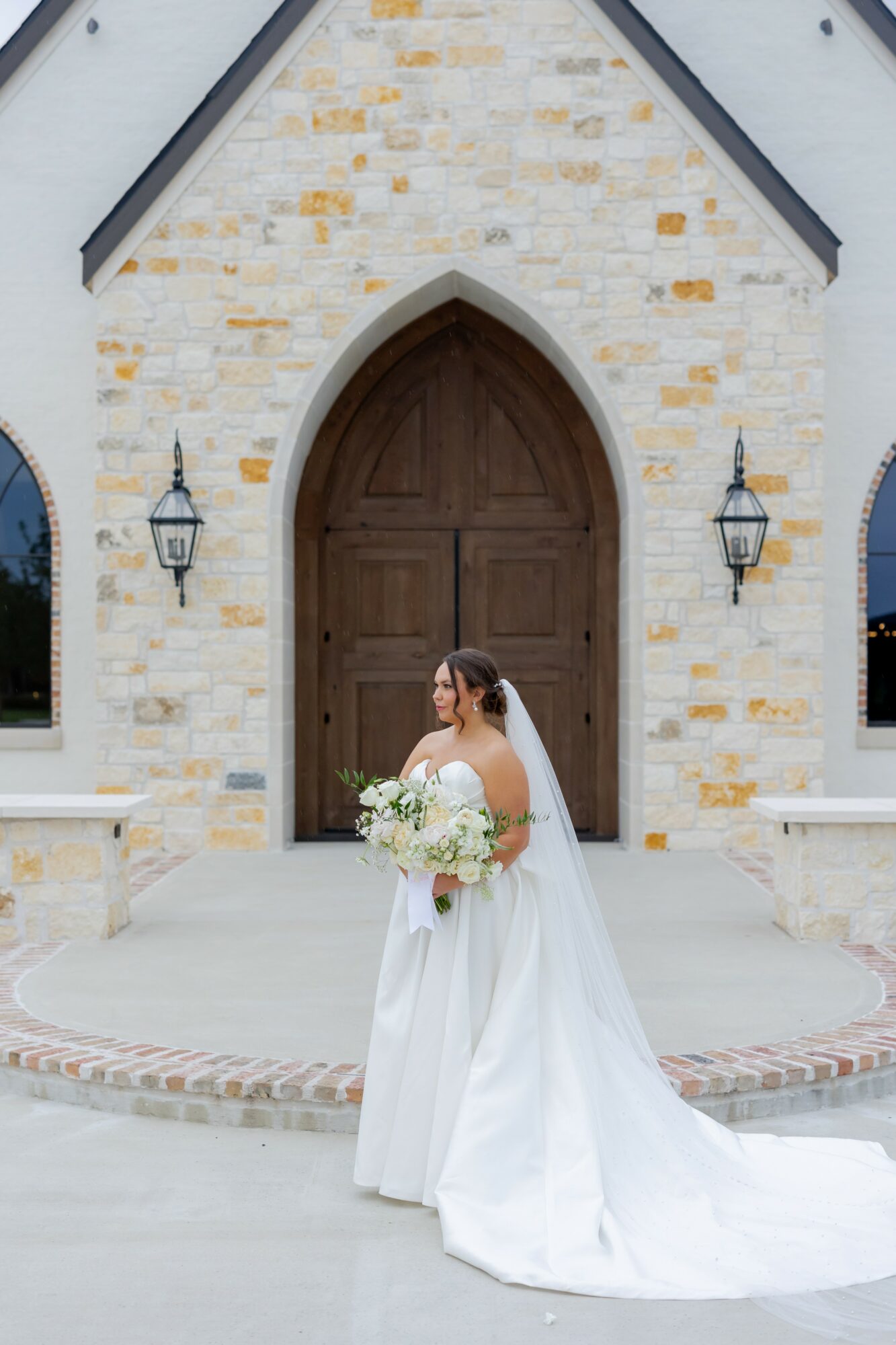 Bride in white dress holding a bouquet standing in front of a church entrance with wooden door and lanterns