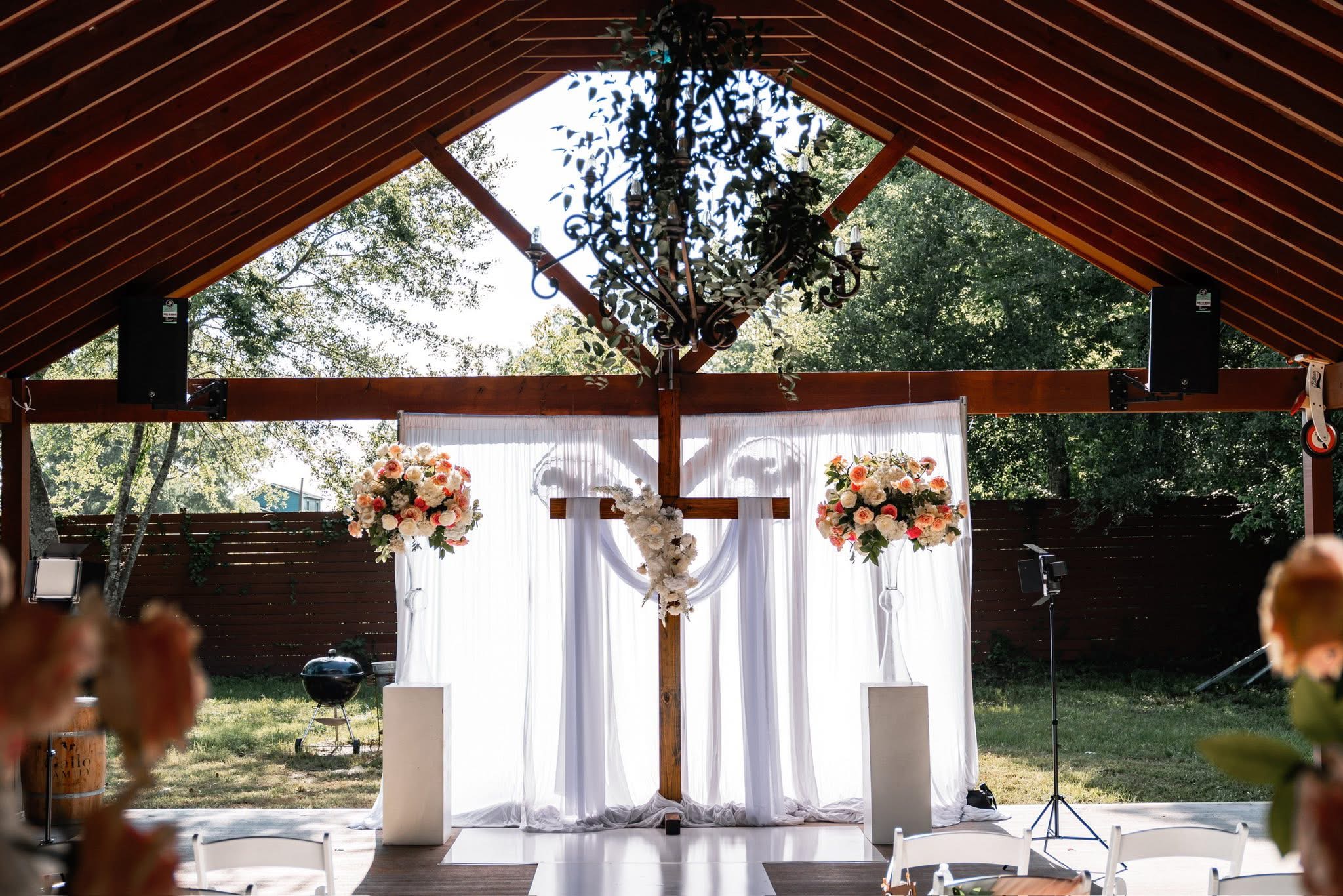 Outdoor wedding altar with white drapes, floral arrangements, and a chandelier under a wooden pavilion, with trees in the background.
