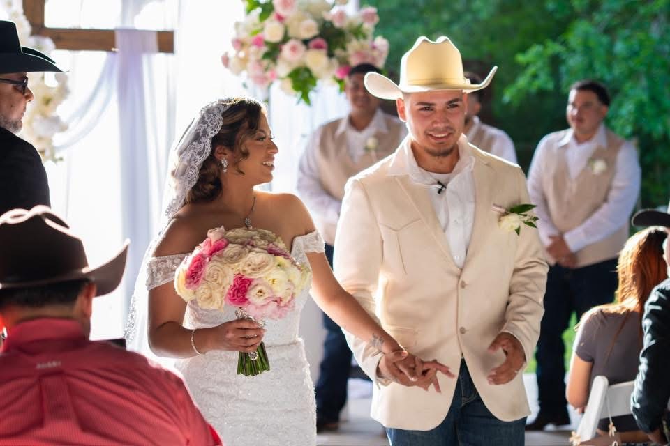 Bride and groom holding hands during wedding ceremony, smiling, with guests and floral decorations in background.
