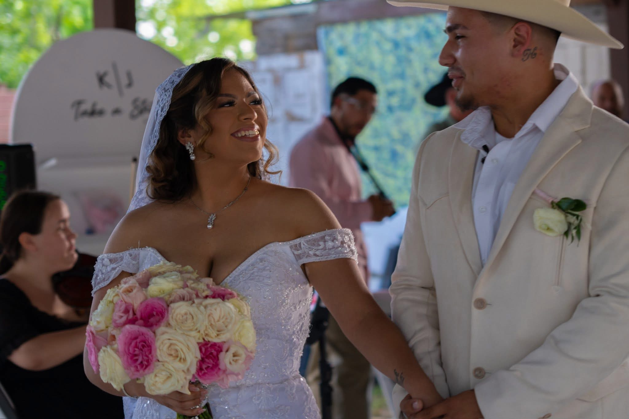 A bride and groom holding hands and smiling at each other during a wedding ceremony, with guests in the background.