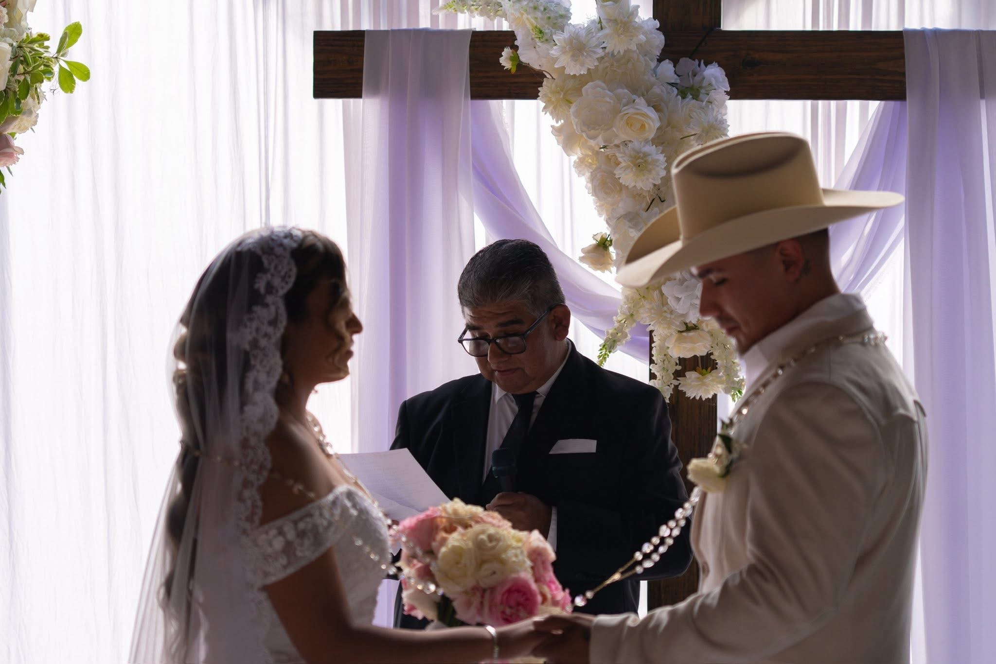 Bride and groom facing each other during wedding ceremony, officiant standing behind them, decorated with white flowers and drapery