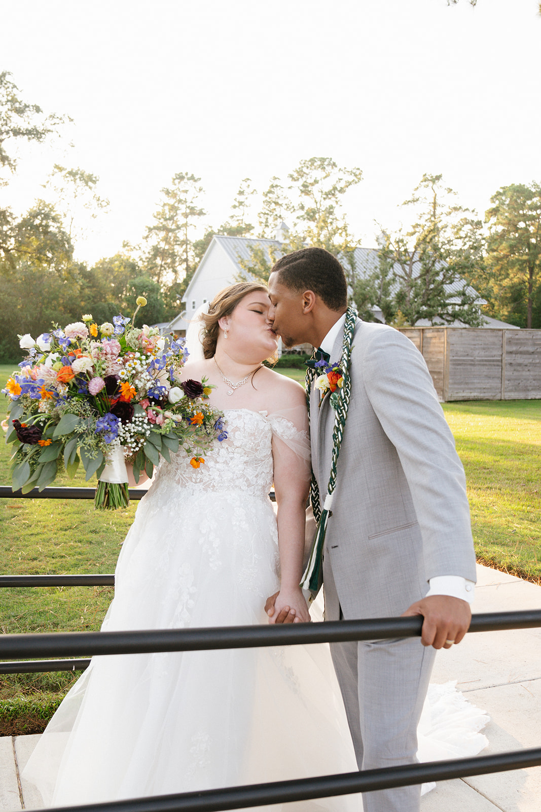 Bride and groom kiss outdoors, bride in white wedding dress holding a colorful bouquet, groom in light gray suit, trees in background.