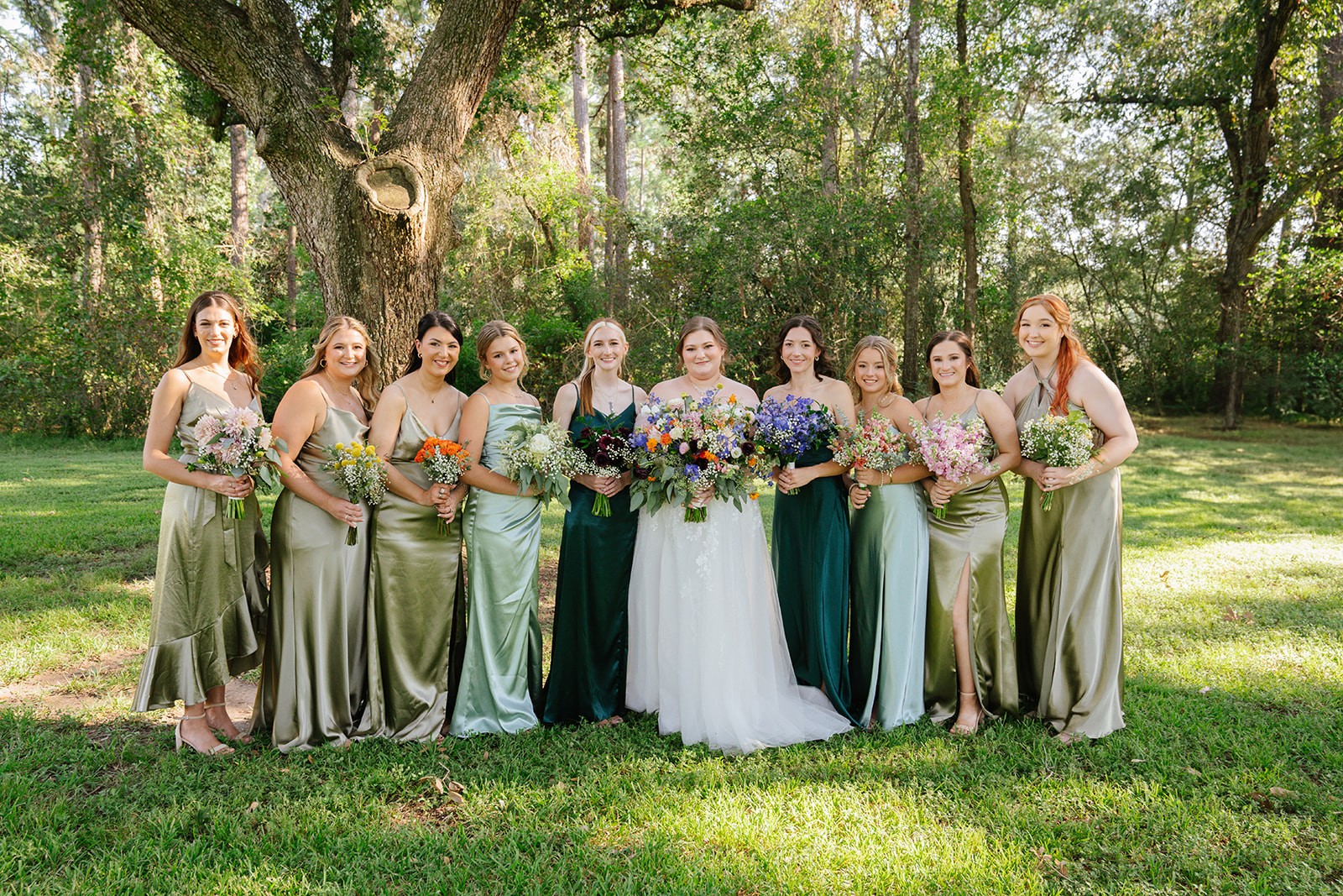 Group of nine women in formal dresses standing outdoors on grass, holding bouquets, with trees in background.