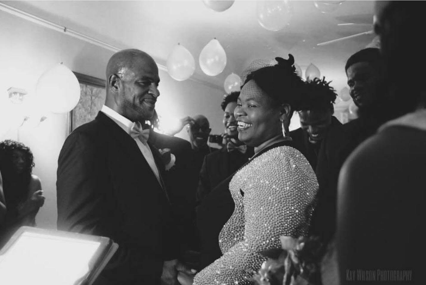 Two people smiling and dancing at a celebration, surrounded by others, with hanging decorations in the background.