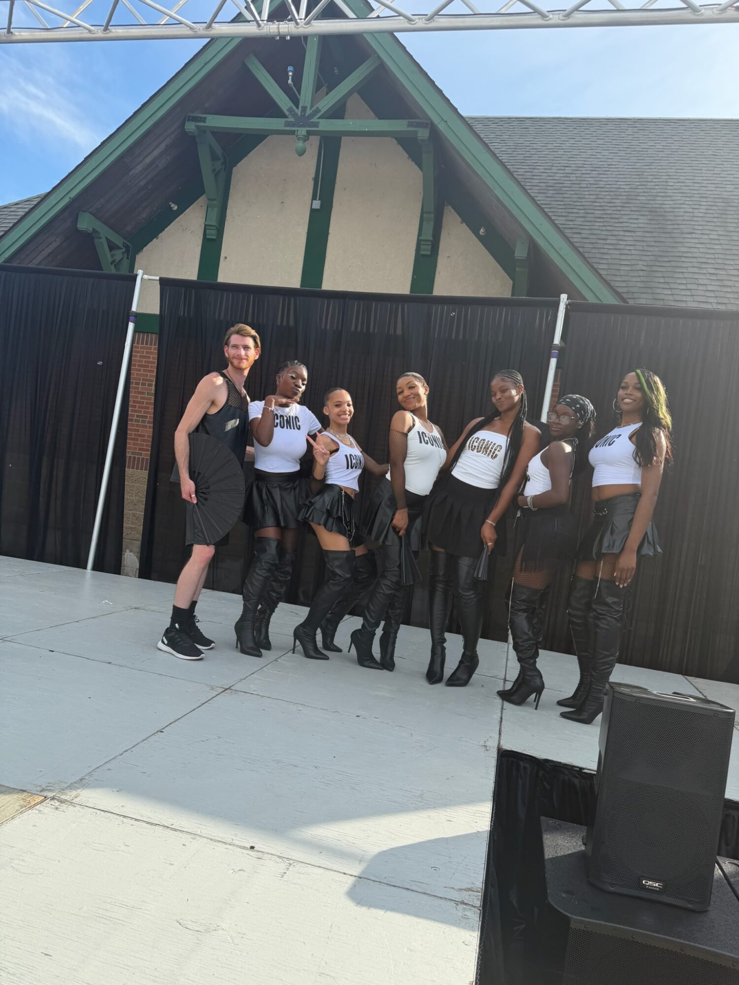 Group of seven women in matching outfits standing outdoors in front of a black fence, with a building in the background.
