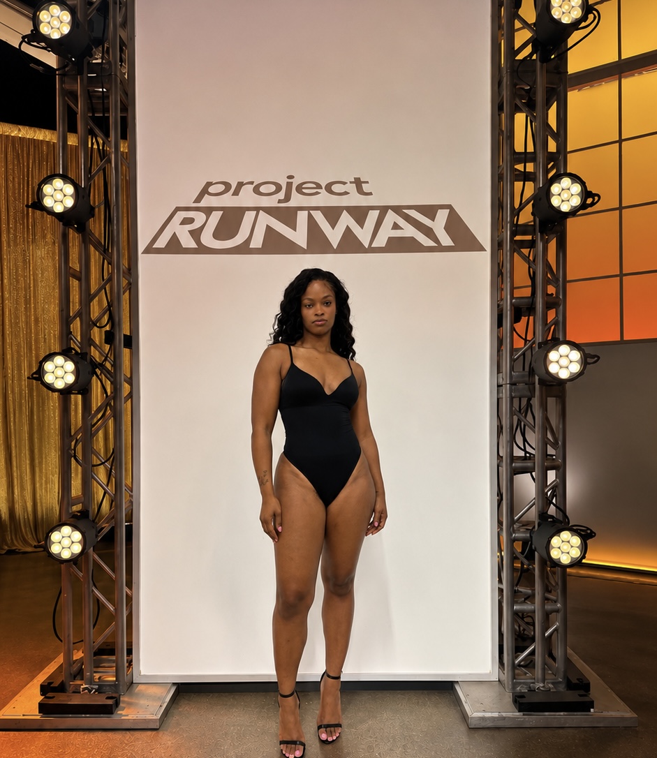 Woman in black swimsuit standing on runway stage with project RUNWAY logo behind her, stage lighting on sides, indoor setting.