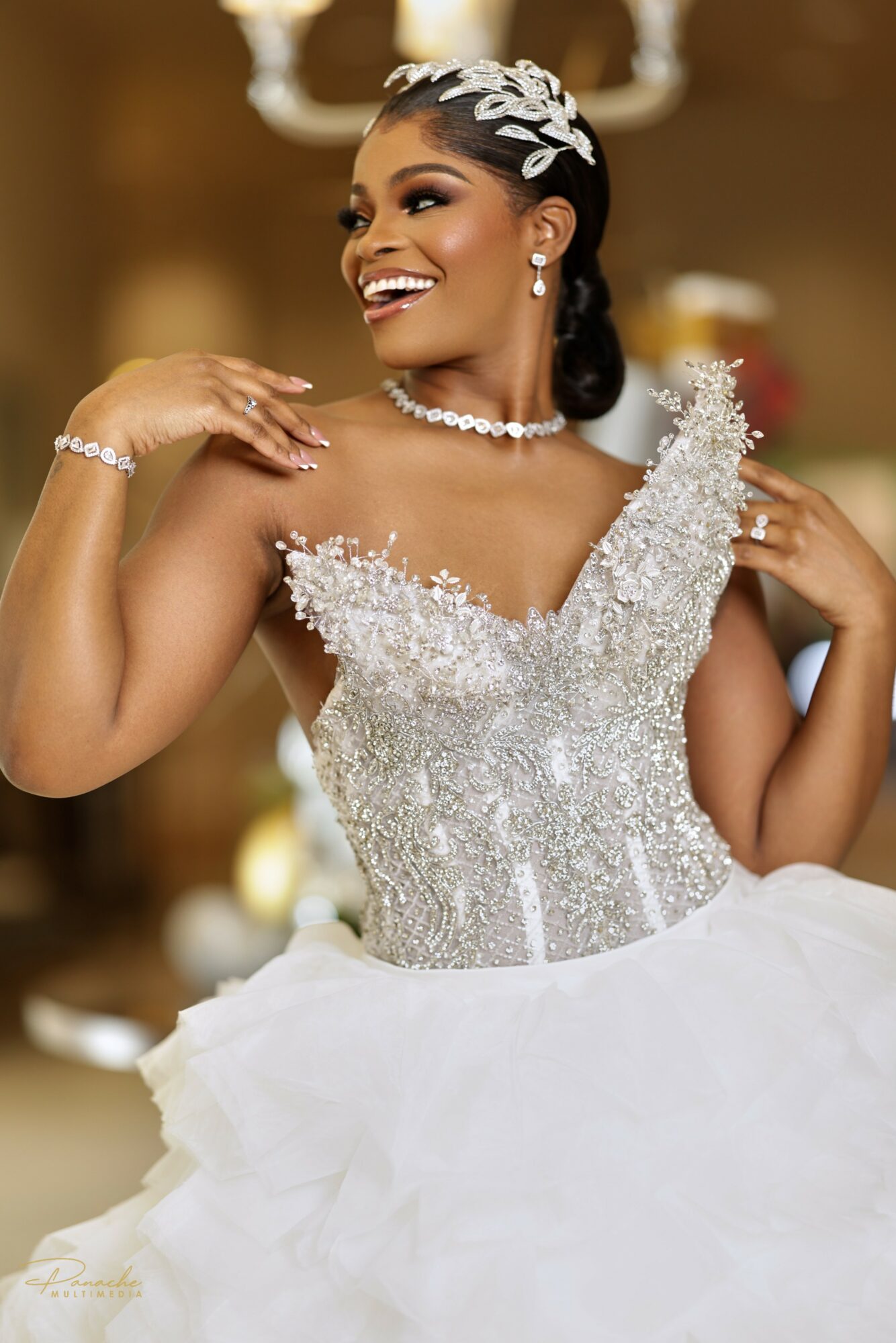 Woman in wedding dress smiling, adjusting her shoulder, with elegant jewelry and a floral headpiece, indoors.