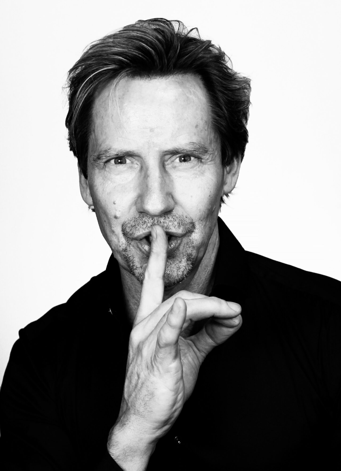 Man with tousled hair and beard holding finger to lips in a shushing gesture, black and white photo.
