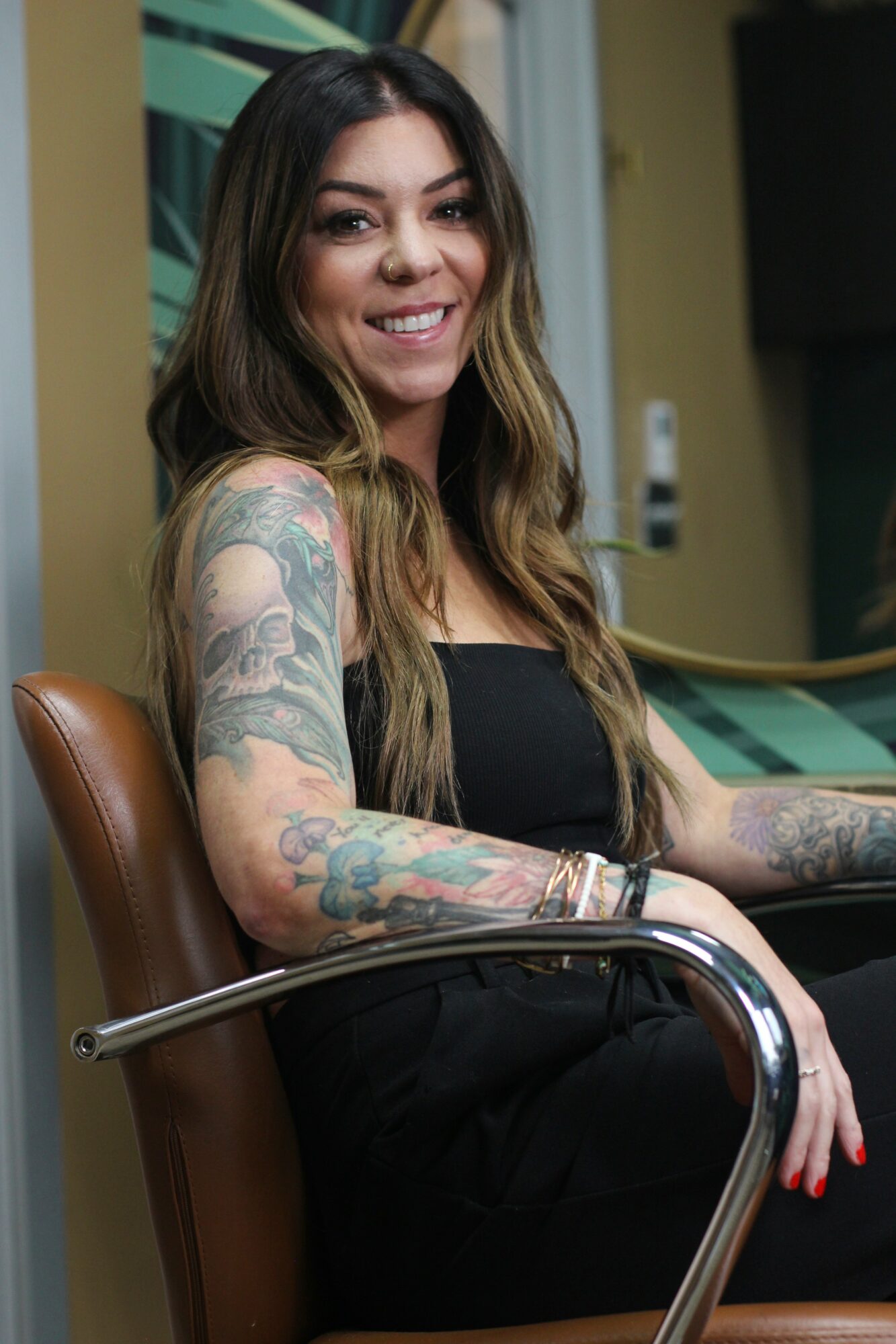 Smiling woman with long hair and tattoos on arms, sitting on a chair indoors.