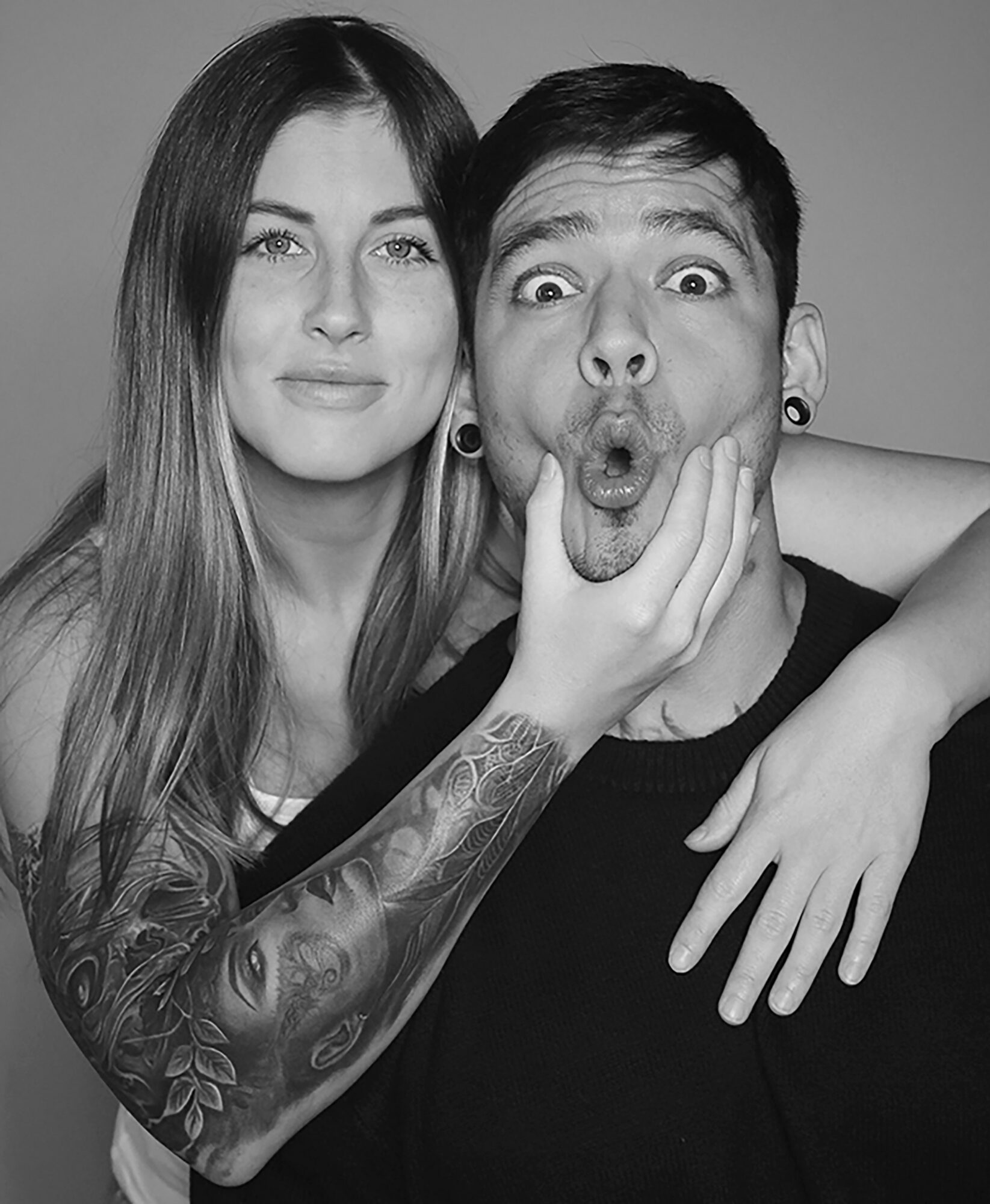 Two people, one with long hair and tattoos, making a surprised face, with the other person smiling beside them.