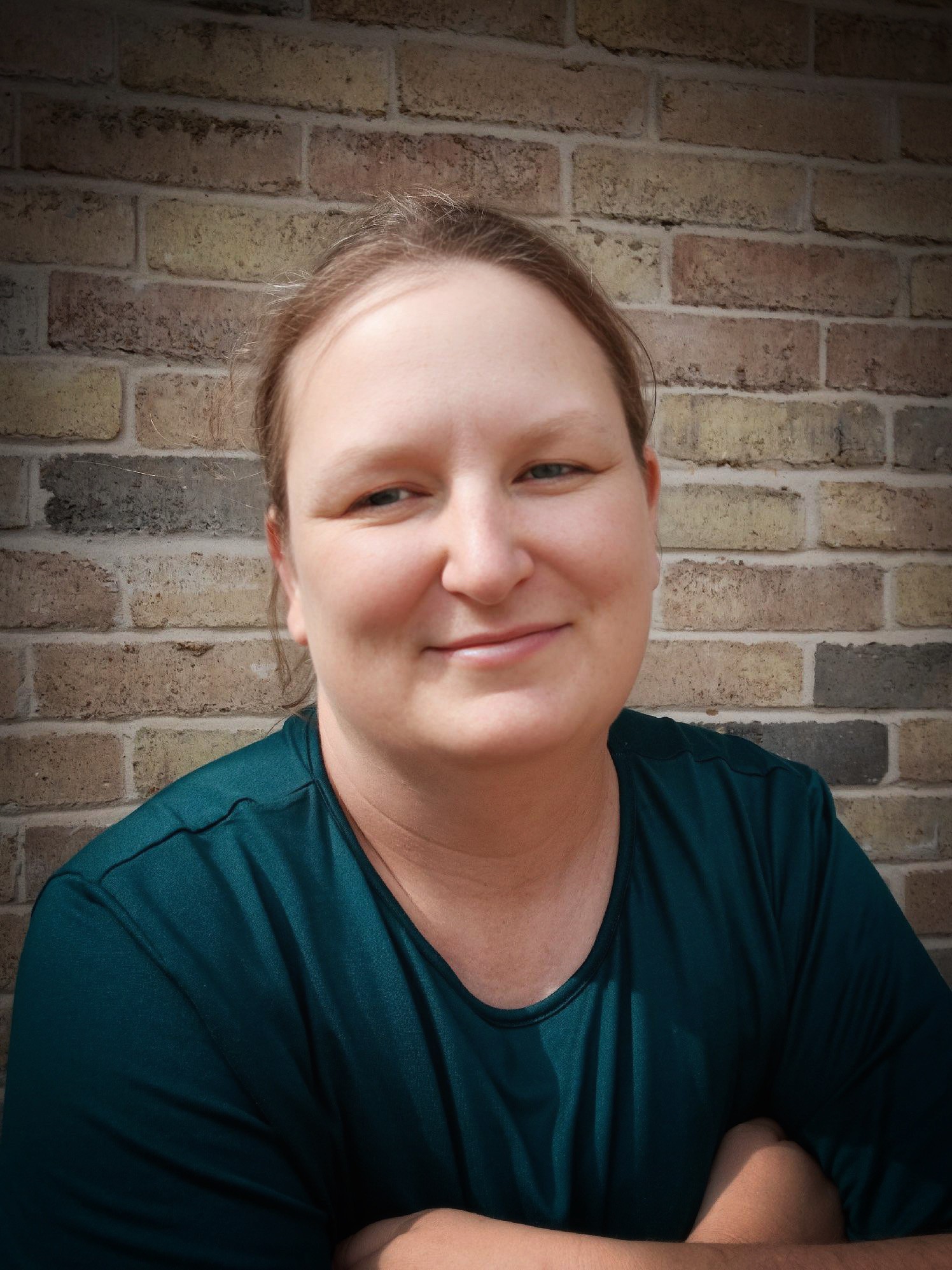 Woman with light brown hair smiling, wearing a dark teal shirt, standing in front of a brick wall.