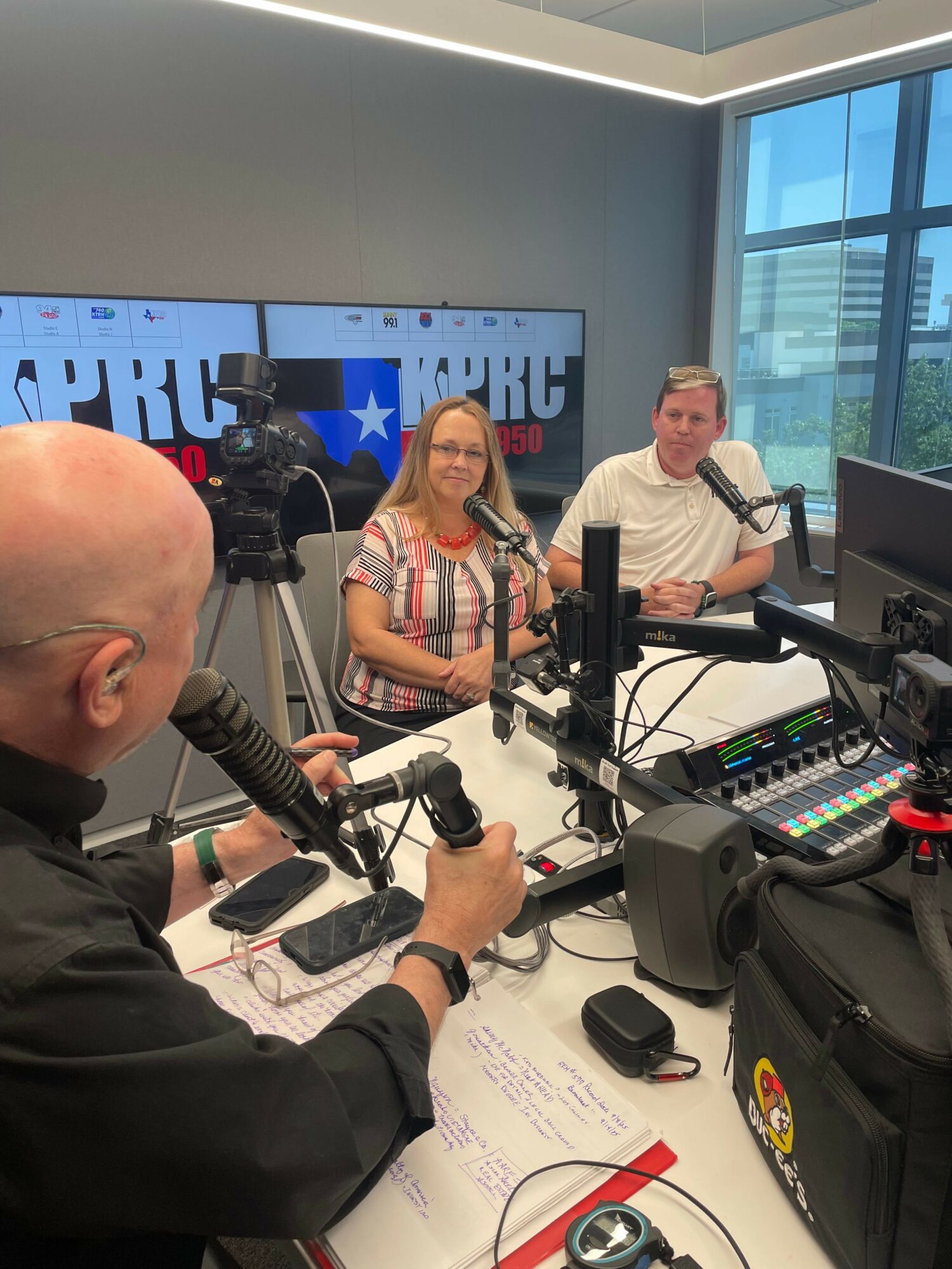 Two women and a man in a radio studio with microphones and equipment, one woman speaking into a microphone.