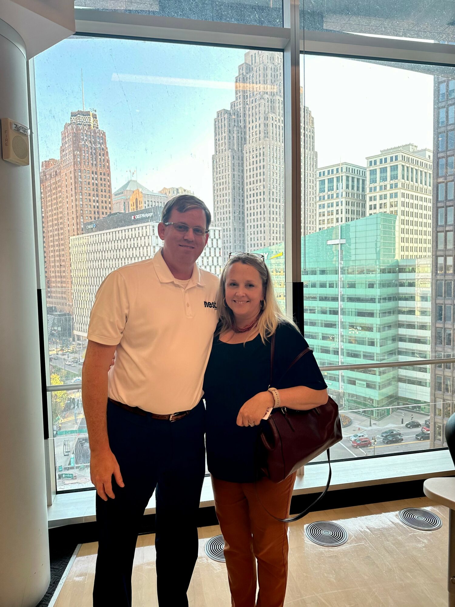 A man and woman stand together indoors with a cityscape view through large windows behind them.