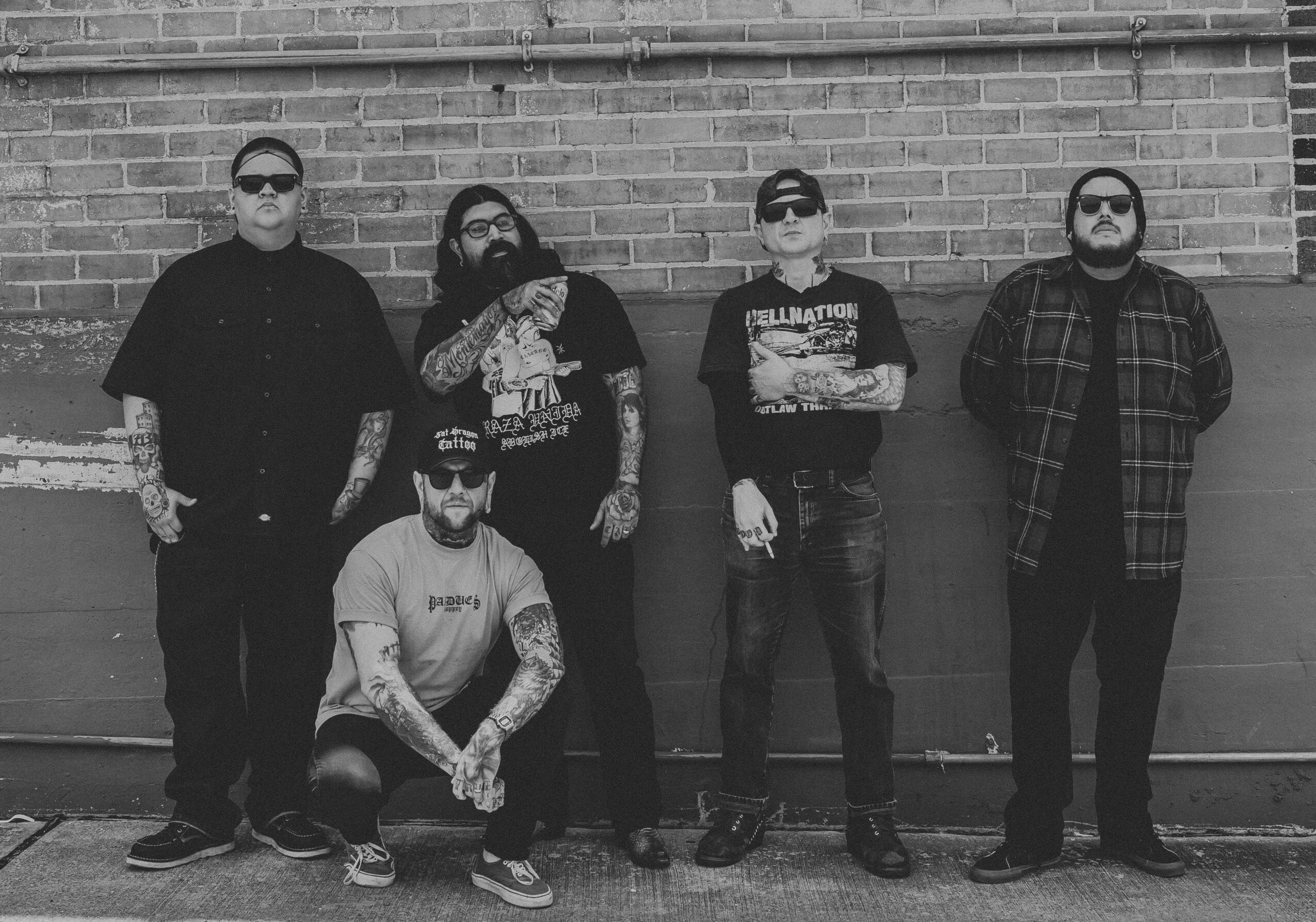 Group of five men standing against a brick wall, one kneeling in front, all wearing casual clothing and sunglasses.