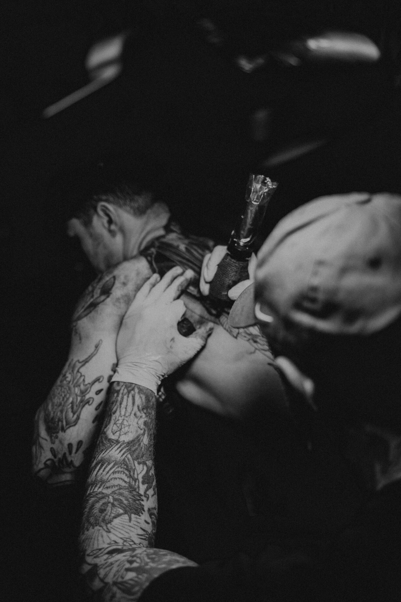 Person receiving a tattoo on their shoulder, with tattoo artist working on them, in a dark setting.