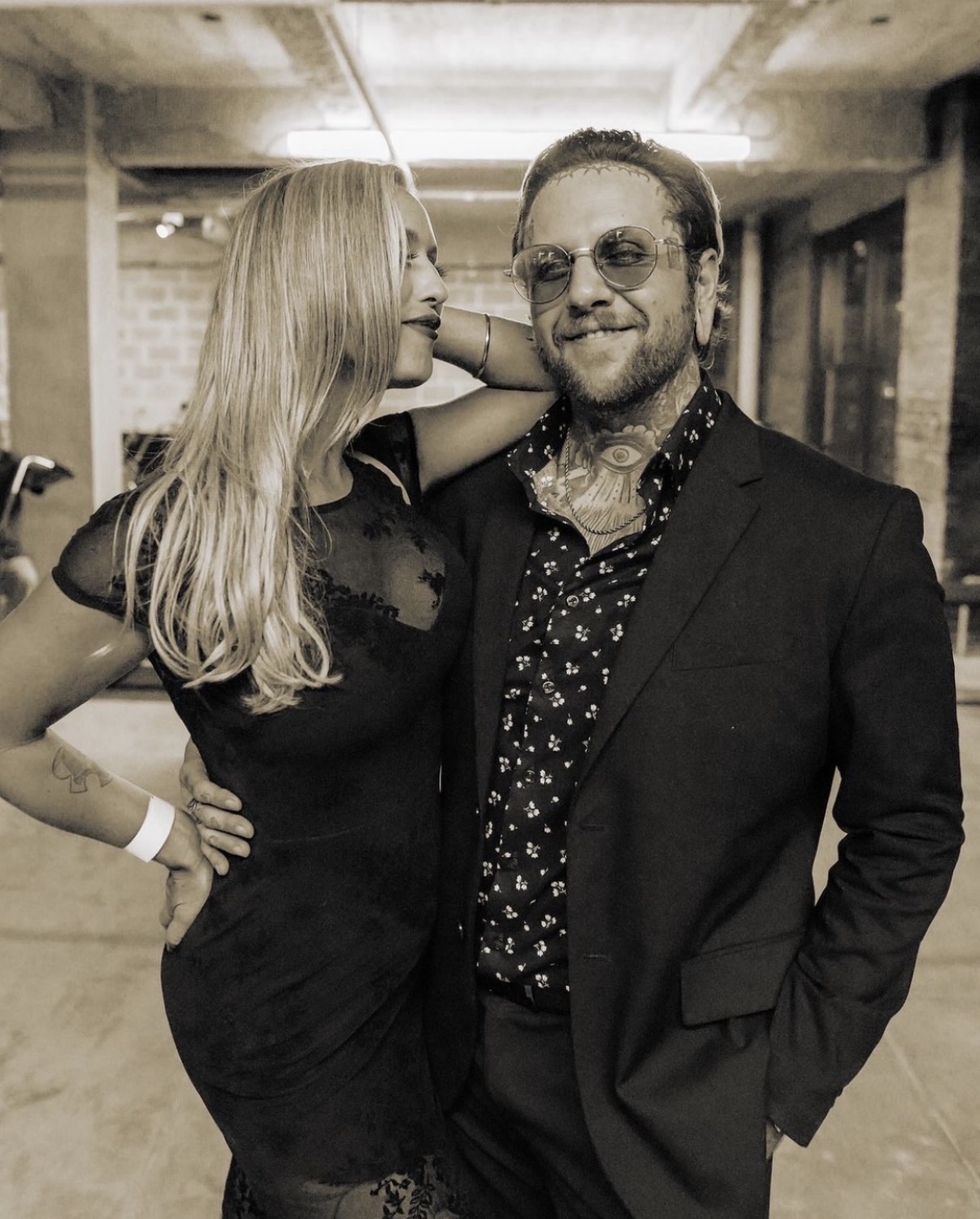A woman and a man pose together indoors, smiling, with the woman leaning toward the man and the man wearing glasses.