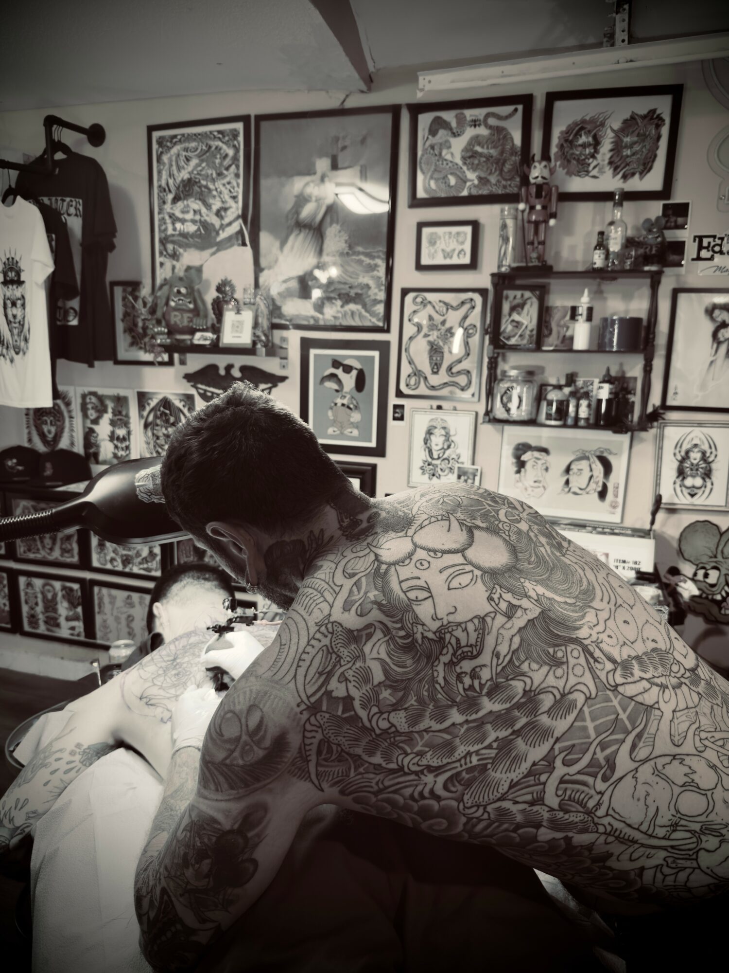 Person with tattoos leaning over a person in a tattoo parlor, surrounded by framed artwork and shelves with supplies.