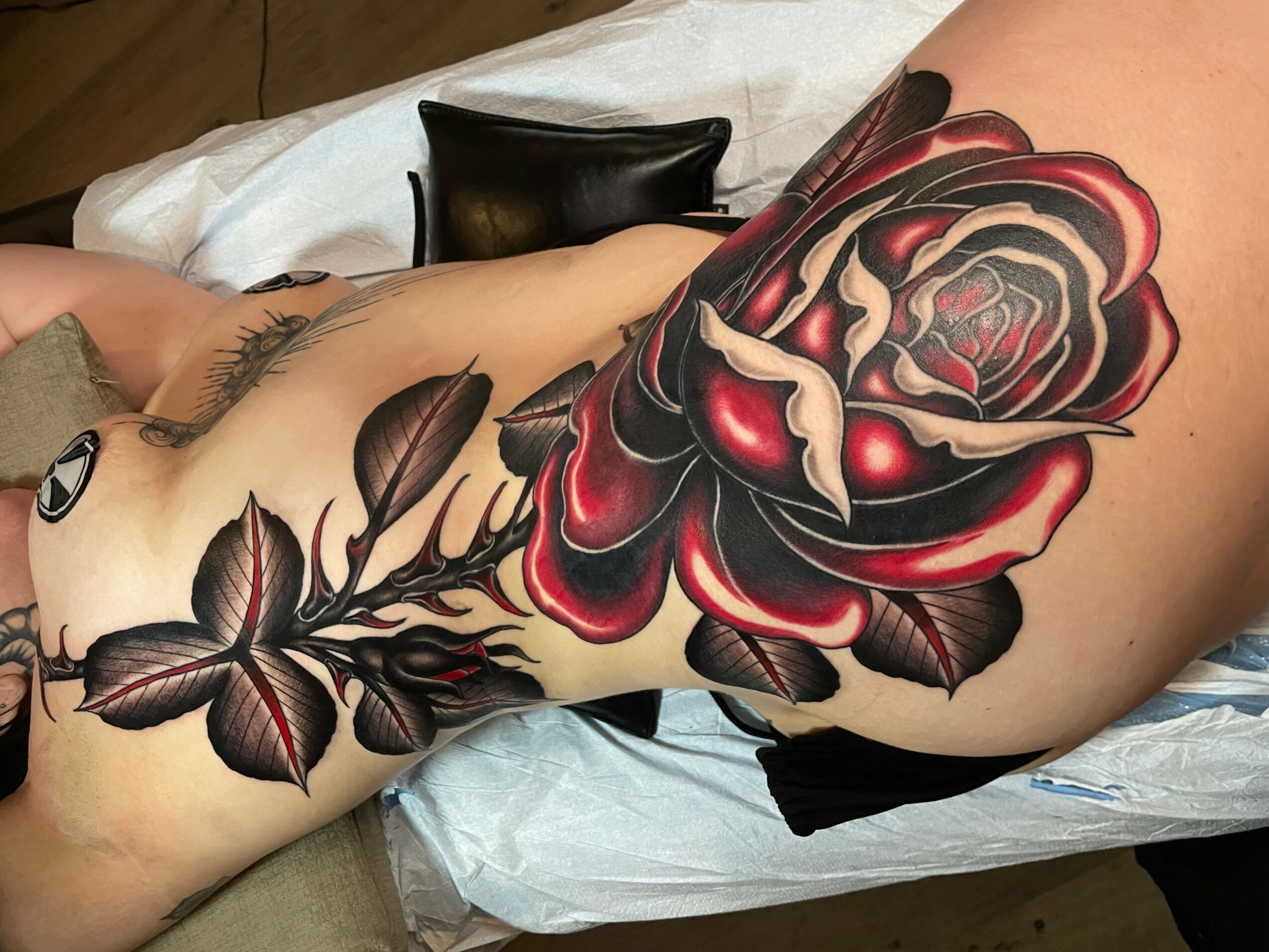 Person with a large, detailed black and red rose tattoo on their thigh, lying on a bed with a black wallet nearby.