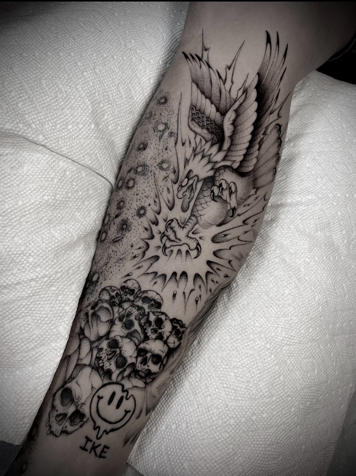 Tattoo of a bird flying above skulls and a smiling face on a person's arm, with detailed shading and line work.