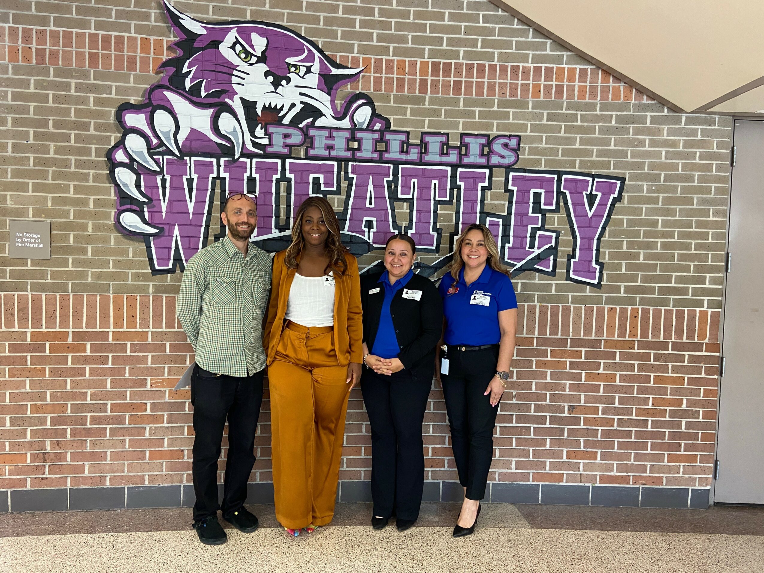 Four people standing in front of a wall with a purple and white Wildcats logo and text, smiling.