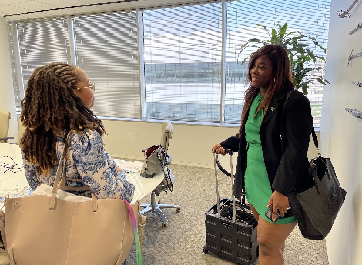 Two women talking in an office with large windows, one with curly hair and the other with straight hair, carrying bags.