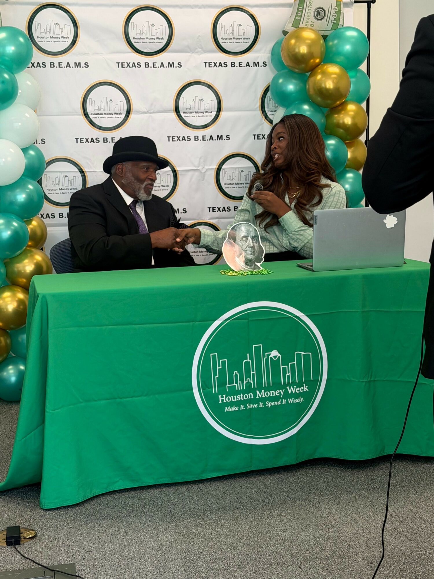 People sitting at a table with a green tablecloth, backdrop with circles and balloons, and a city skyline logo.