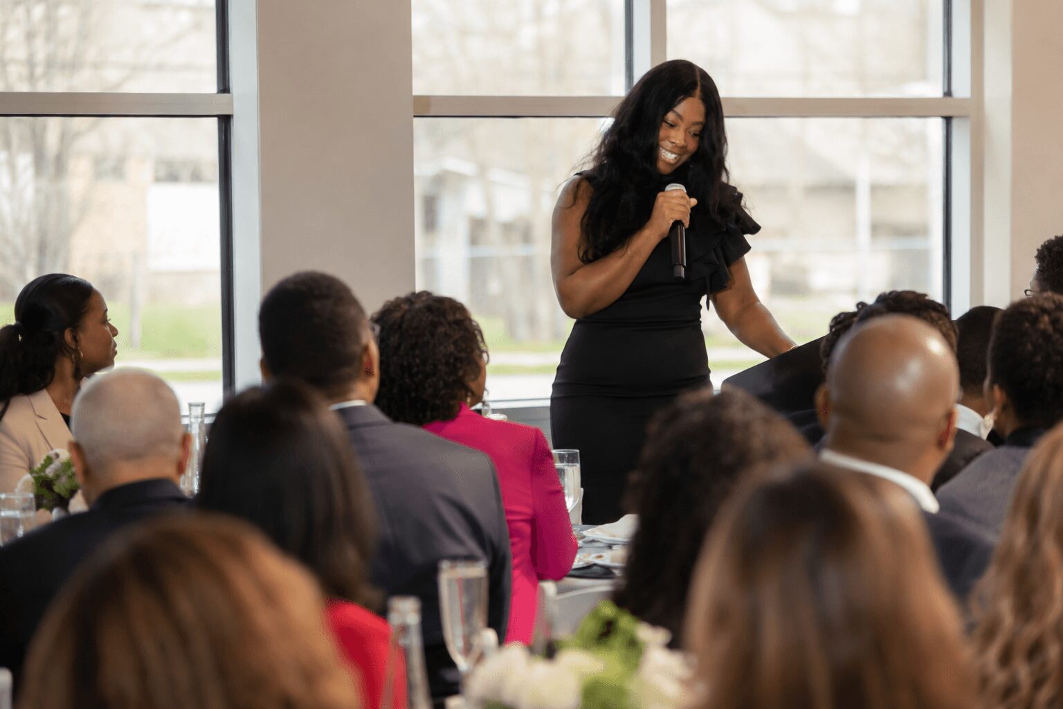 Woman speaking to an audience in a room with large windows, audience members seated and listening attentively.