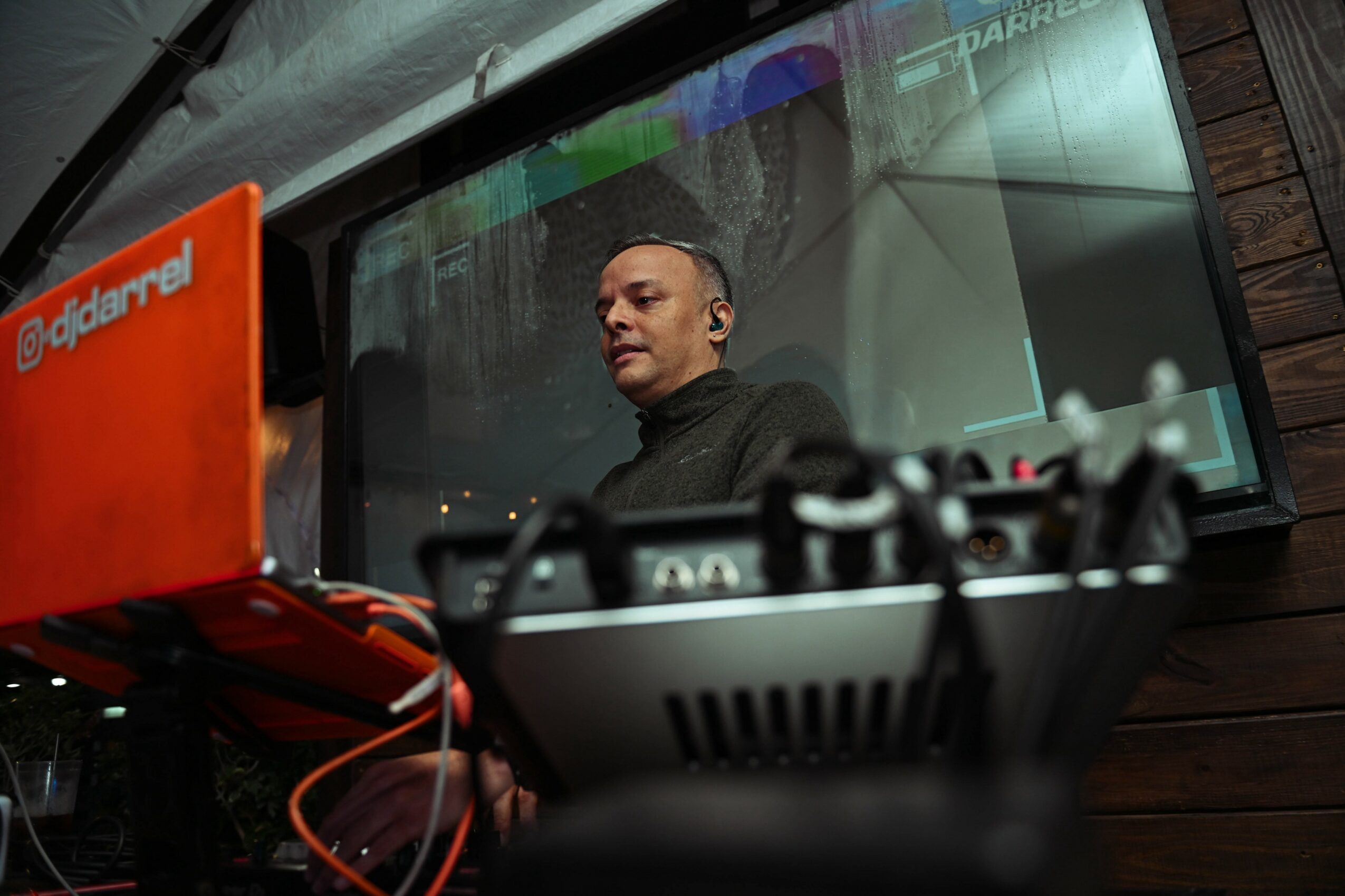 Person with short hair and black shirt looking to the side, behind DJ equipment, in a dimly lit setting.