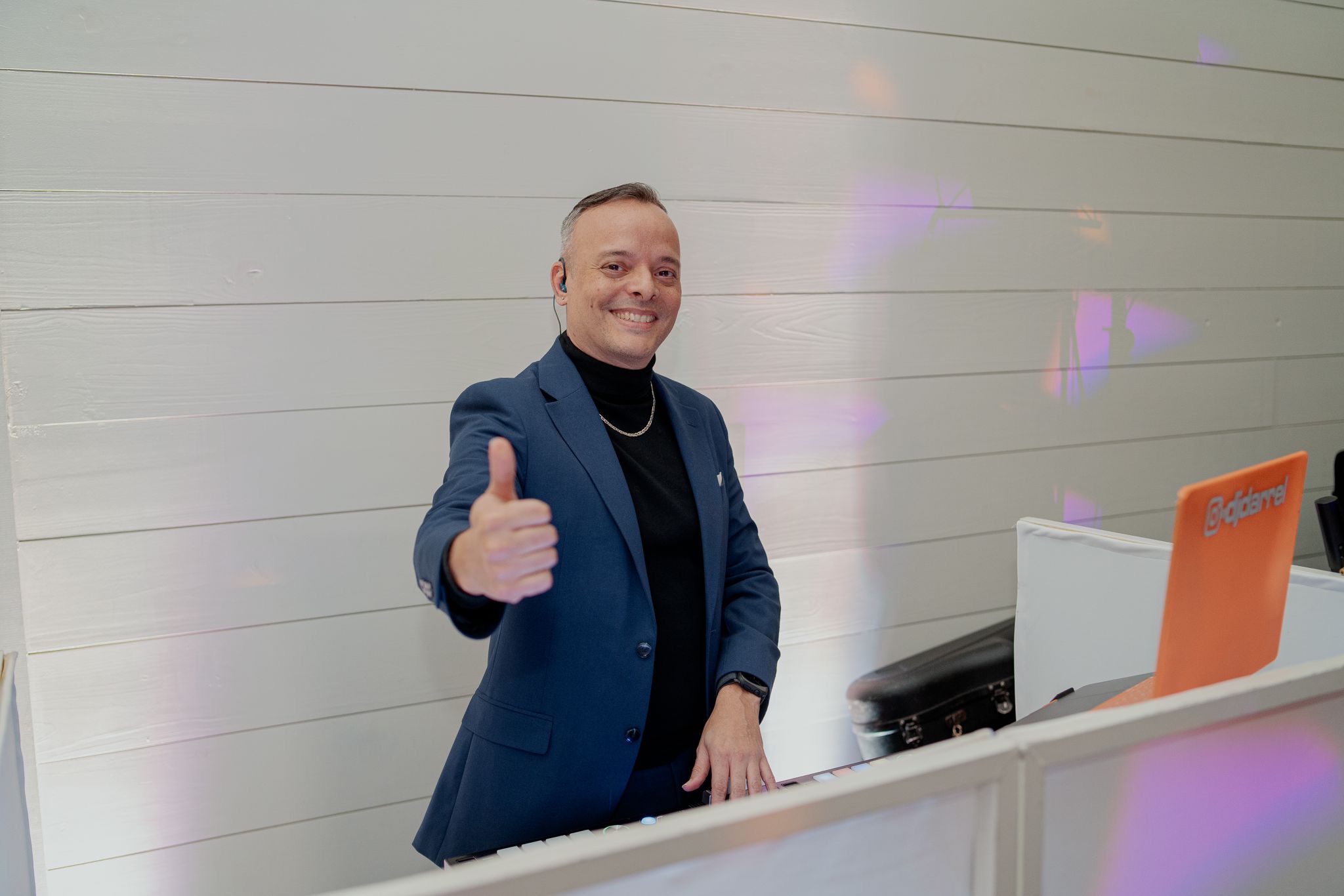 Man in a blue suit giving a thumbs-up, standing behind a white counter with a laptop, smiling.