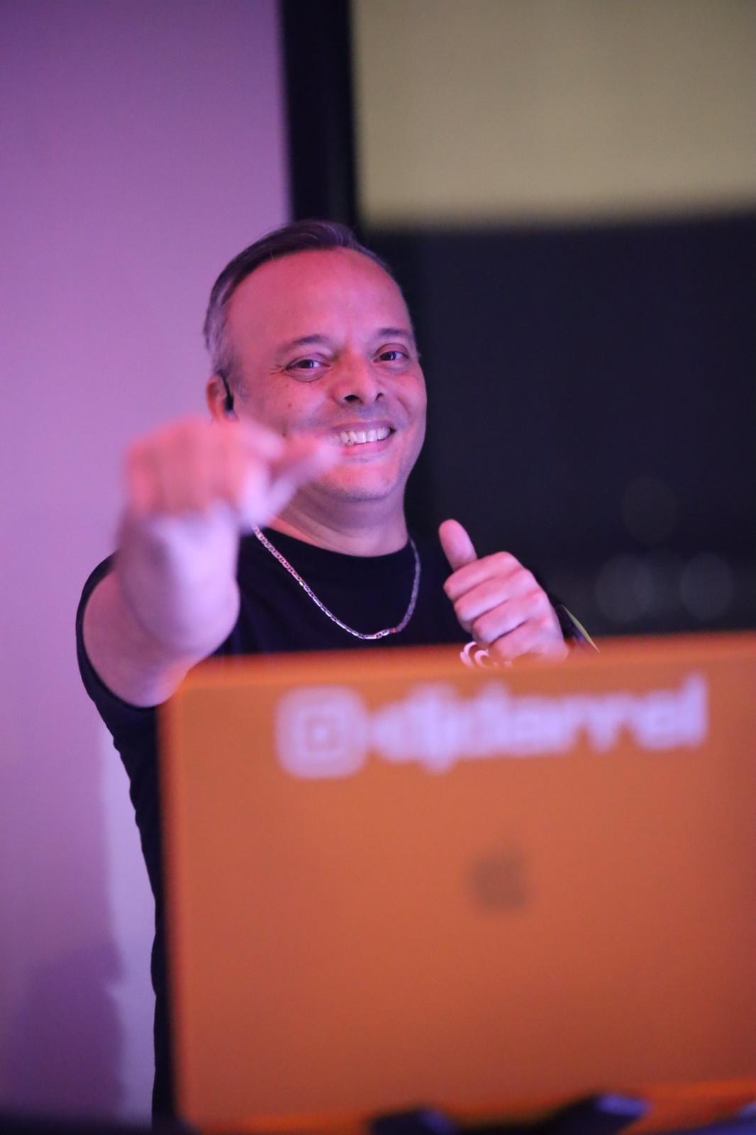 Person smiling and holding a microphone, pointing towards the camera, with a blurred background and a laptop in front.