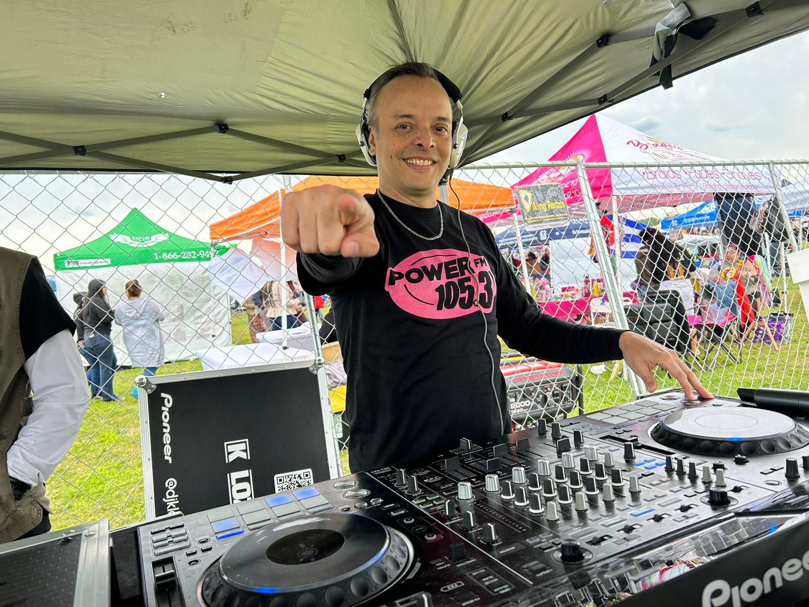 DJ wearing headphones and a black shirt with pink text, pointing at the camera with a smile, at an outdoor event.
